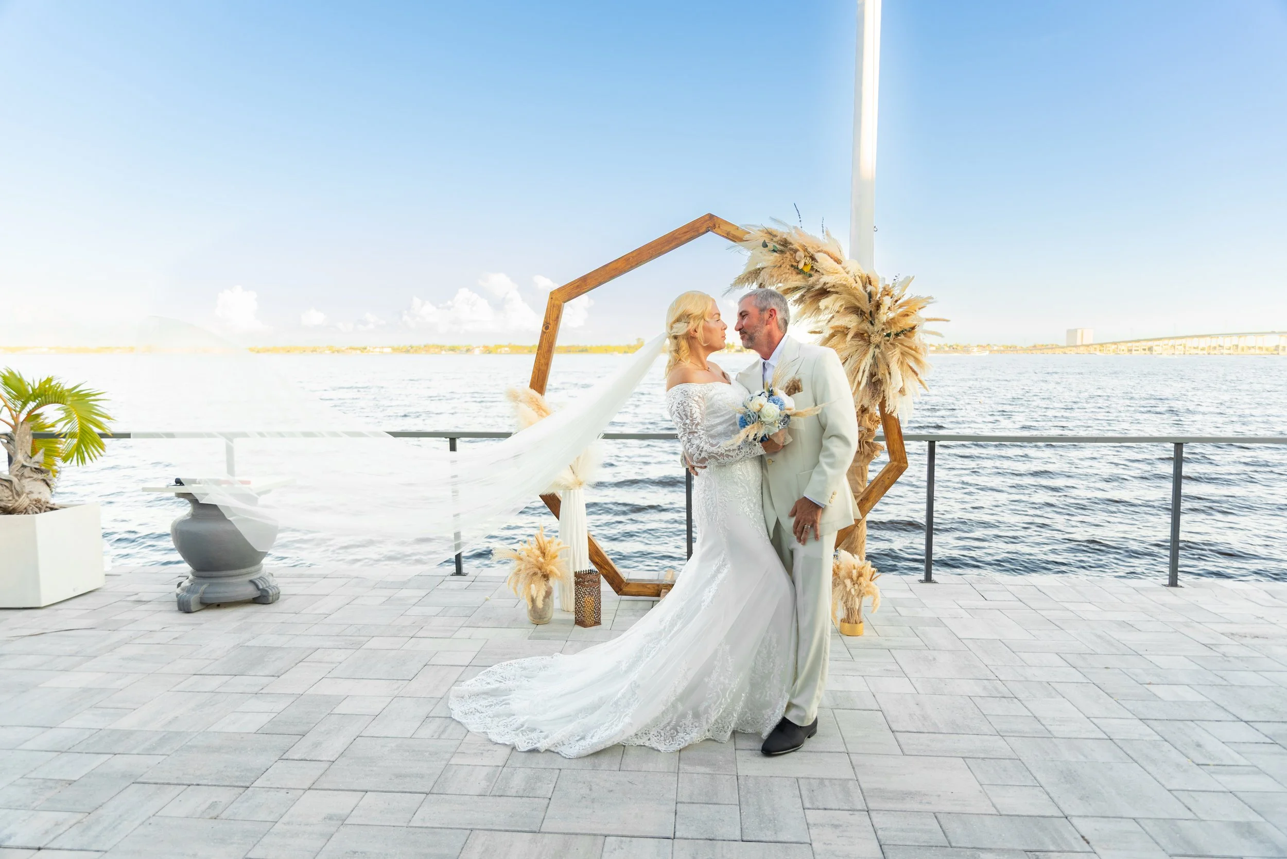 Bride and groom in wedding attire sharing a kiss on a waterfront terrace, with decor including pampas grass, a wooden geometric arch, and potted plants during daytime.