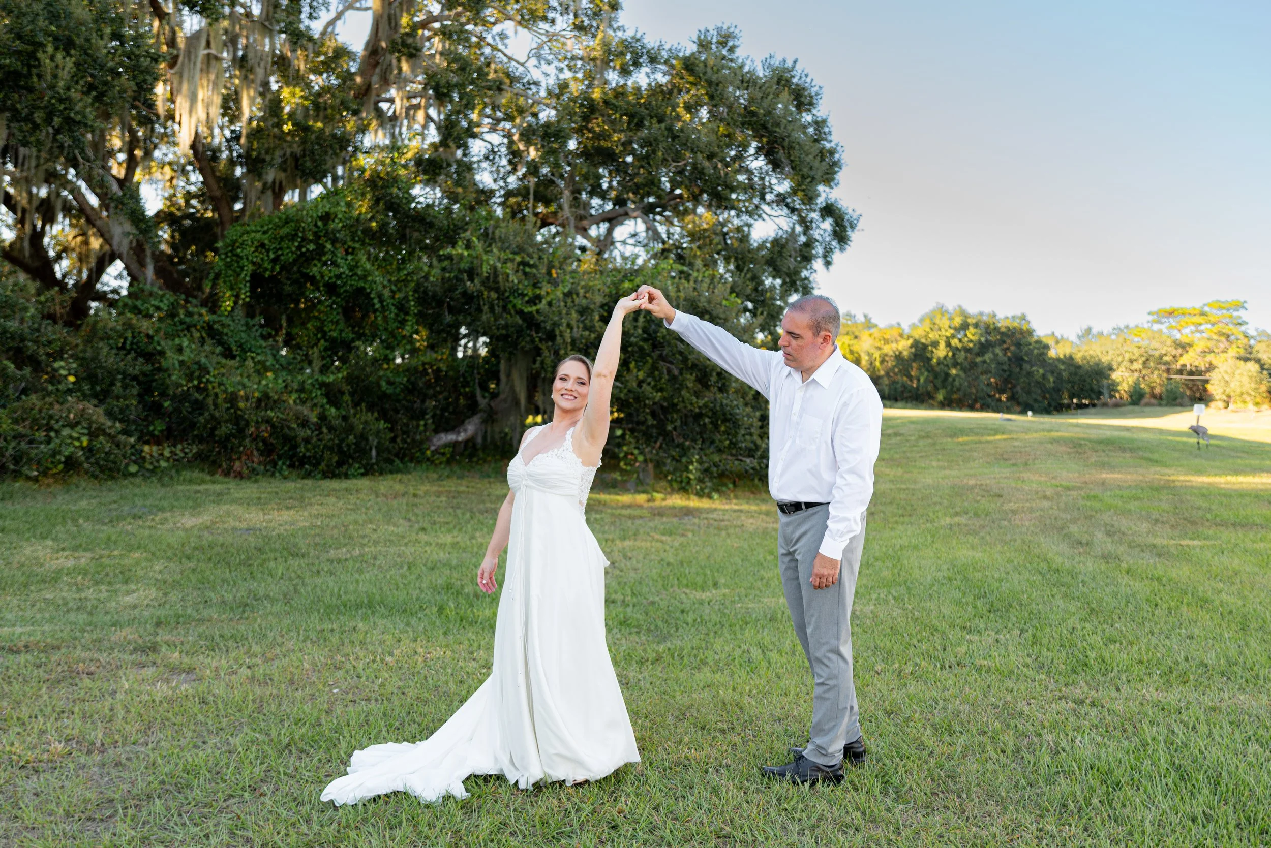 A bride and groom dancing outdoors on a sunny day, with the groom twirling the bride in a grassy park surrounded by trees.