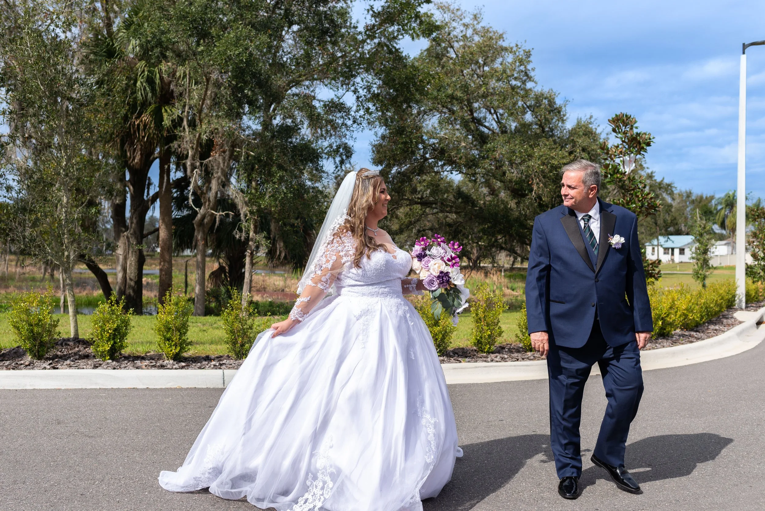 Bride in a wedding dress holding a bouquet of purple and white flowers standing next to a man in a navy suit on a street with trees and a partly cloudy sky.