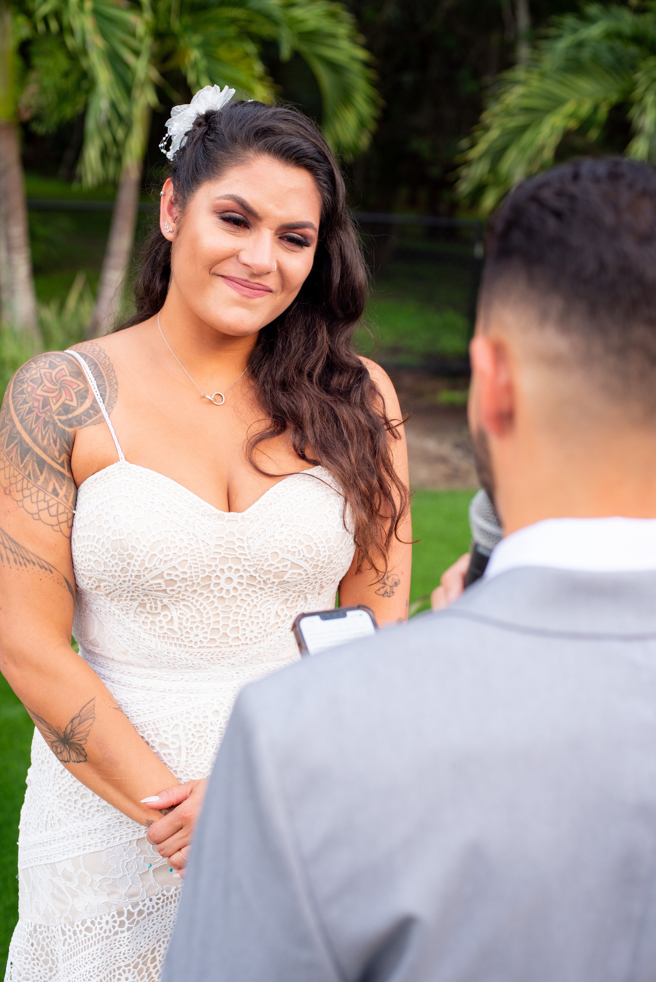 A woman with long wavy dark hair, tattoos on her arms, and wearing a white crochet dress, is smiling as she looks at a man holding a microphone. They are outdoors with green palm trees in the background.