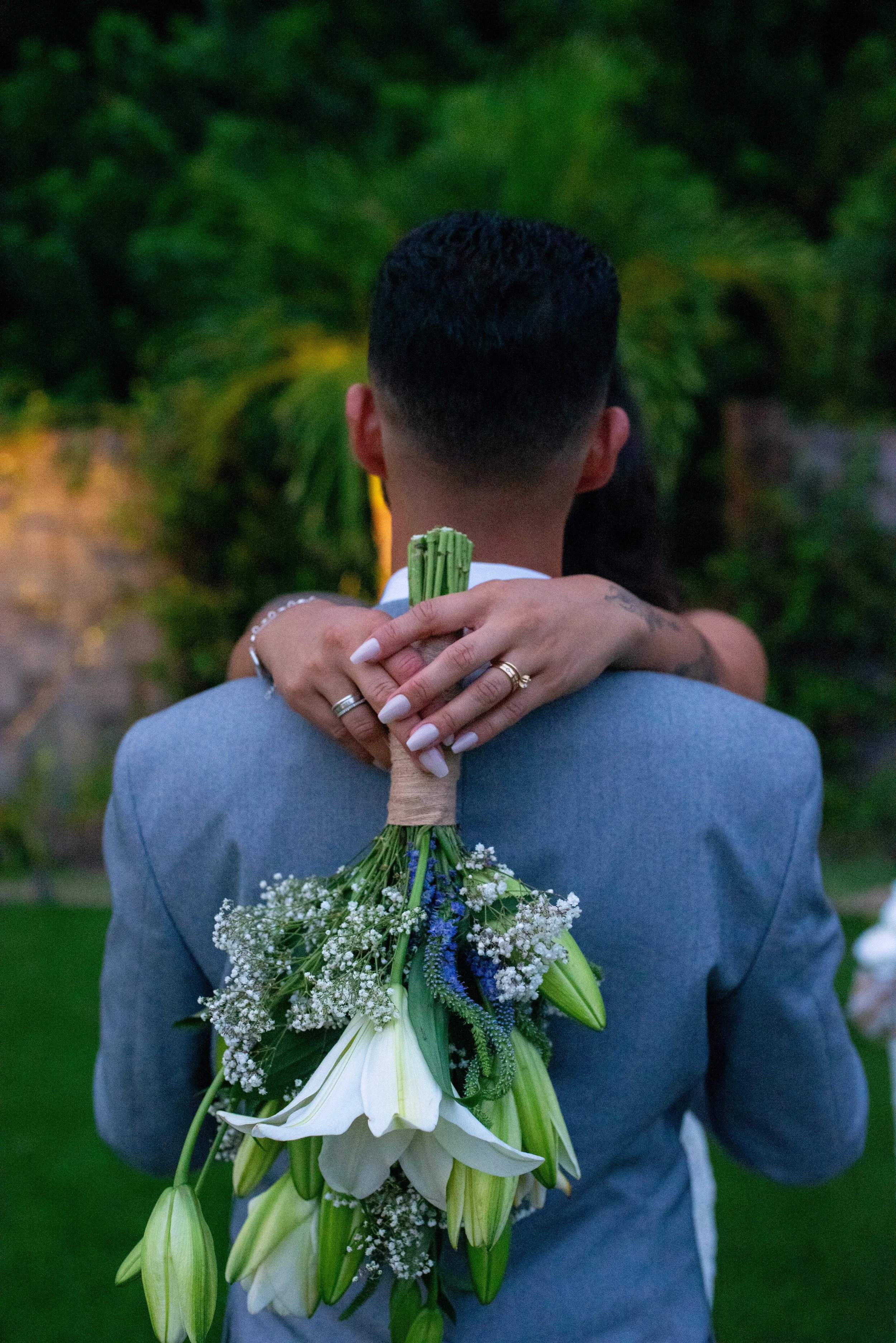 A person wearing a gray suit is holding a bouquet of flowers behind their back, with a woman embracing them from behind, her arms wrapped around their neck. The woman has manicured nails, multiple rings, and tattoos; the bouquet contains white lilies