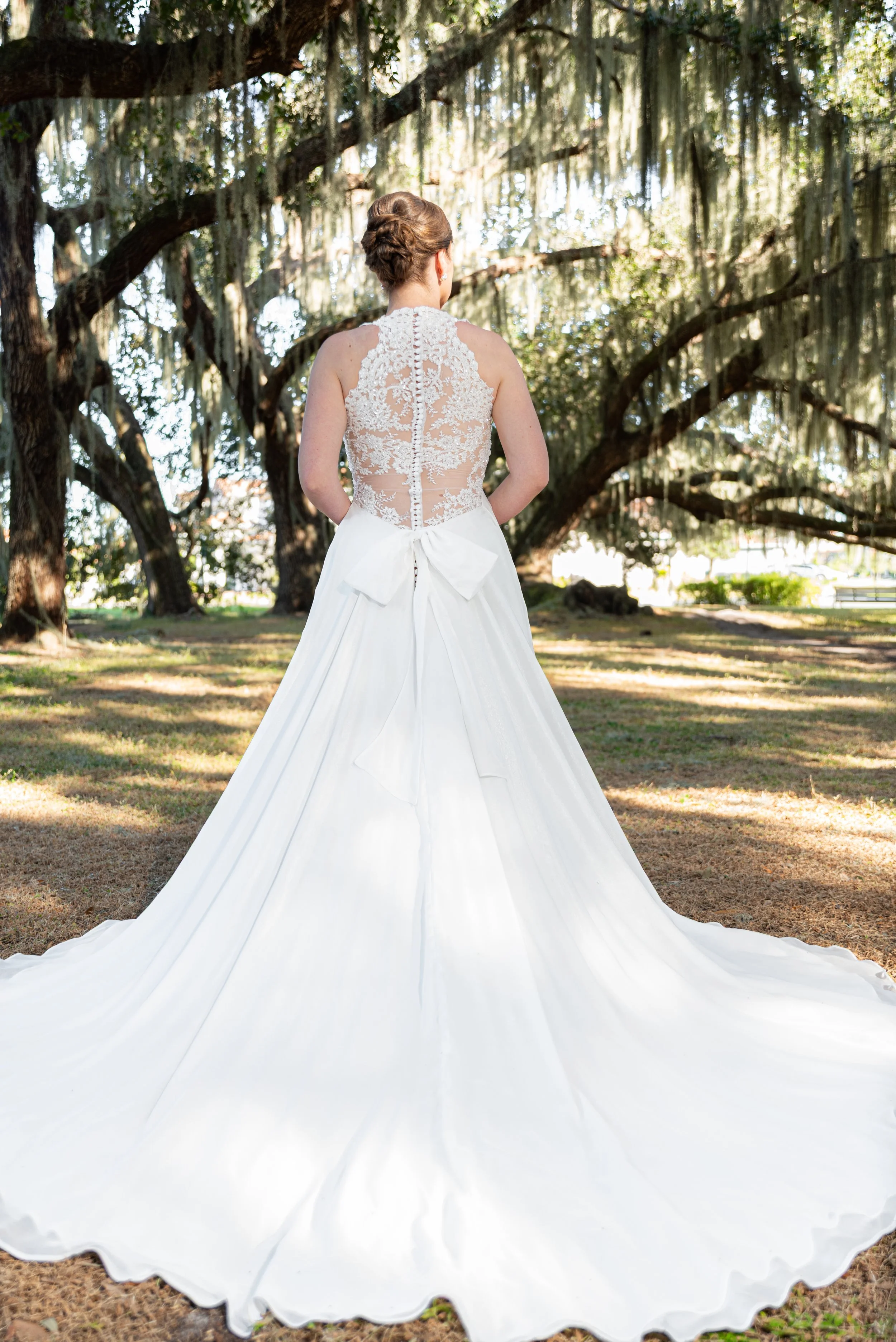 Back view of a bride in a lace and satin wedding dress with a bow, standing outdoors under large trees with hanging moss.