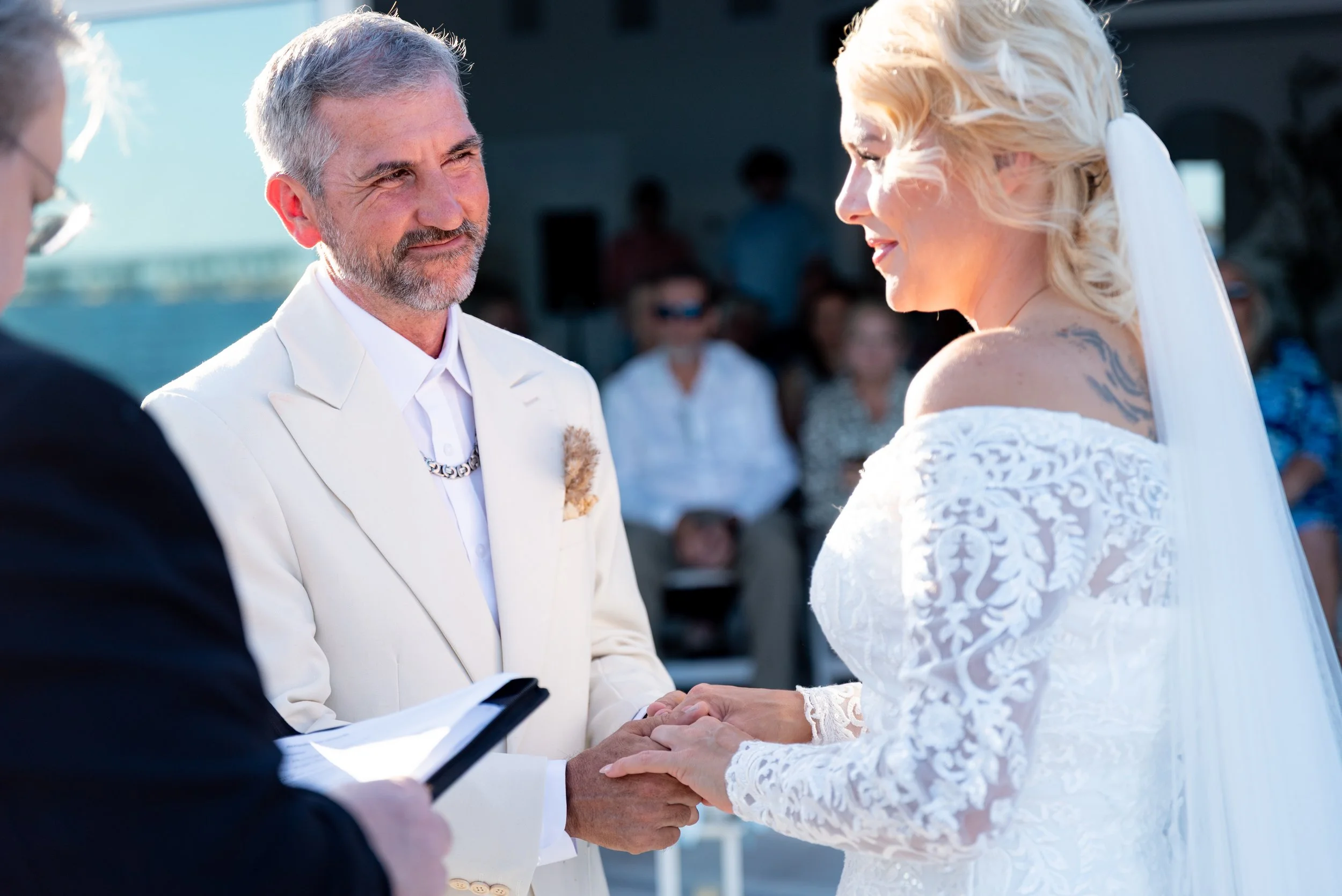 A couple gets married outdoors, exchanging vows and holding hands, with a person officiating the ceremony.