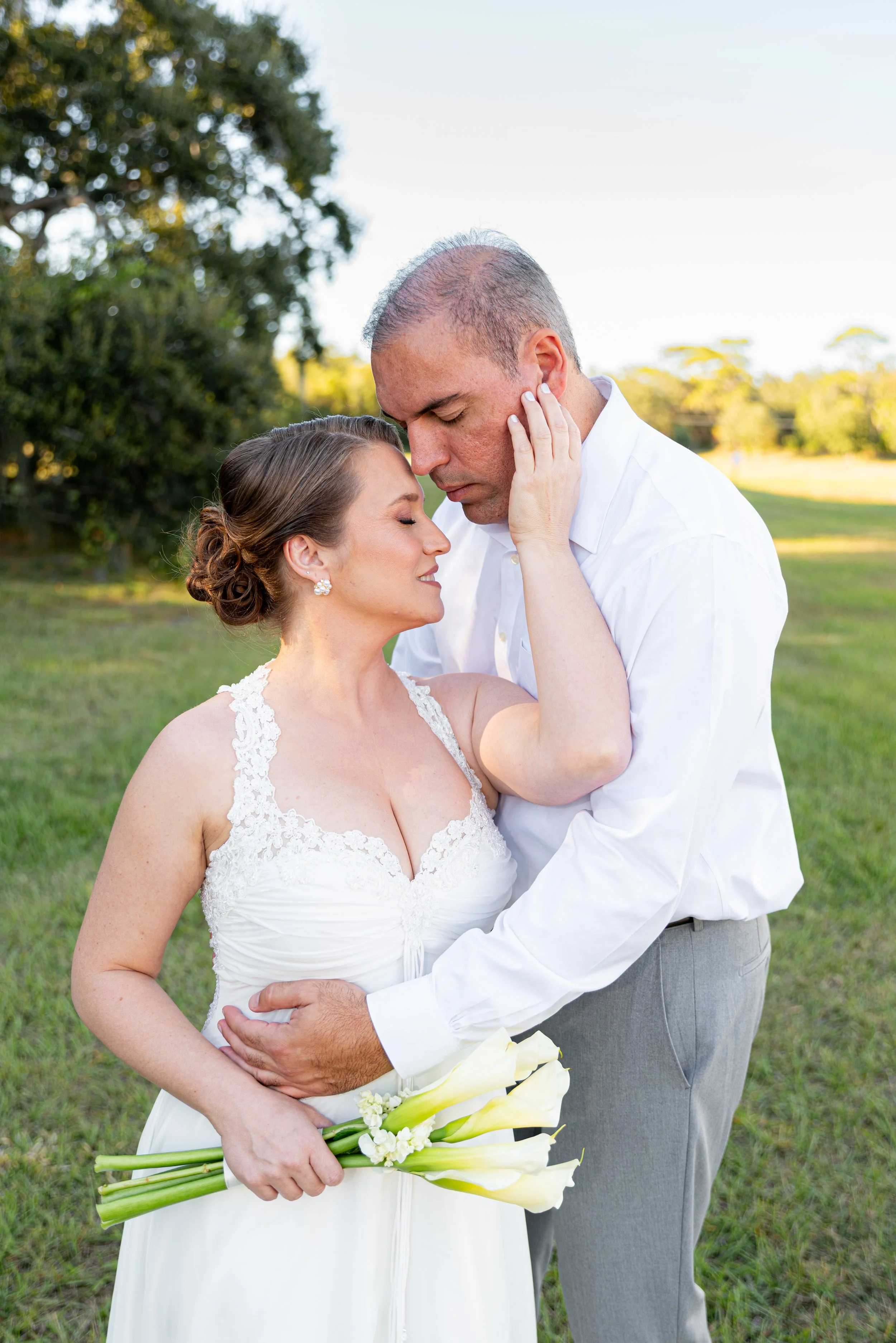 A bride and groom embrace outdoors on their wedding day, with the bride holding a bouquet of white calla lilies.
