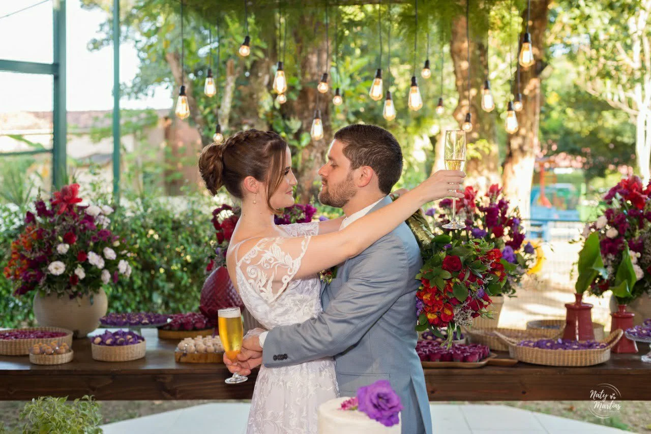A bride and groom sharing a romantic moment with champagne at their wedding reception outdoors, surrounded by flowers and string lights.