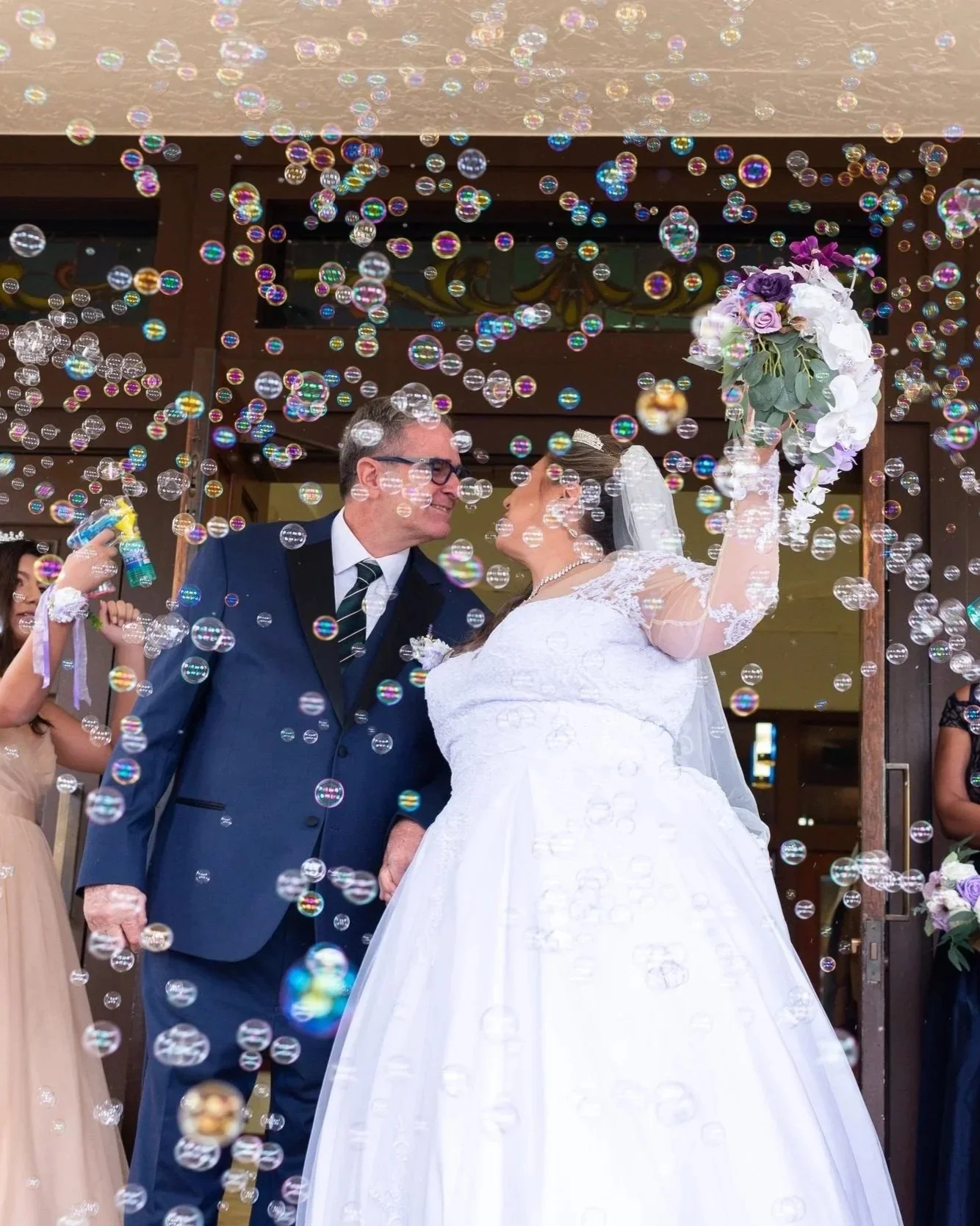 A bride and groom on their wedding day, surrounded by floating bubbles, exchanging a loving look outside a building with wooden doors. The bride is in a white wedding dress holding a bouquet, and the groom is in a blue suit with glasses.