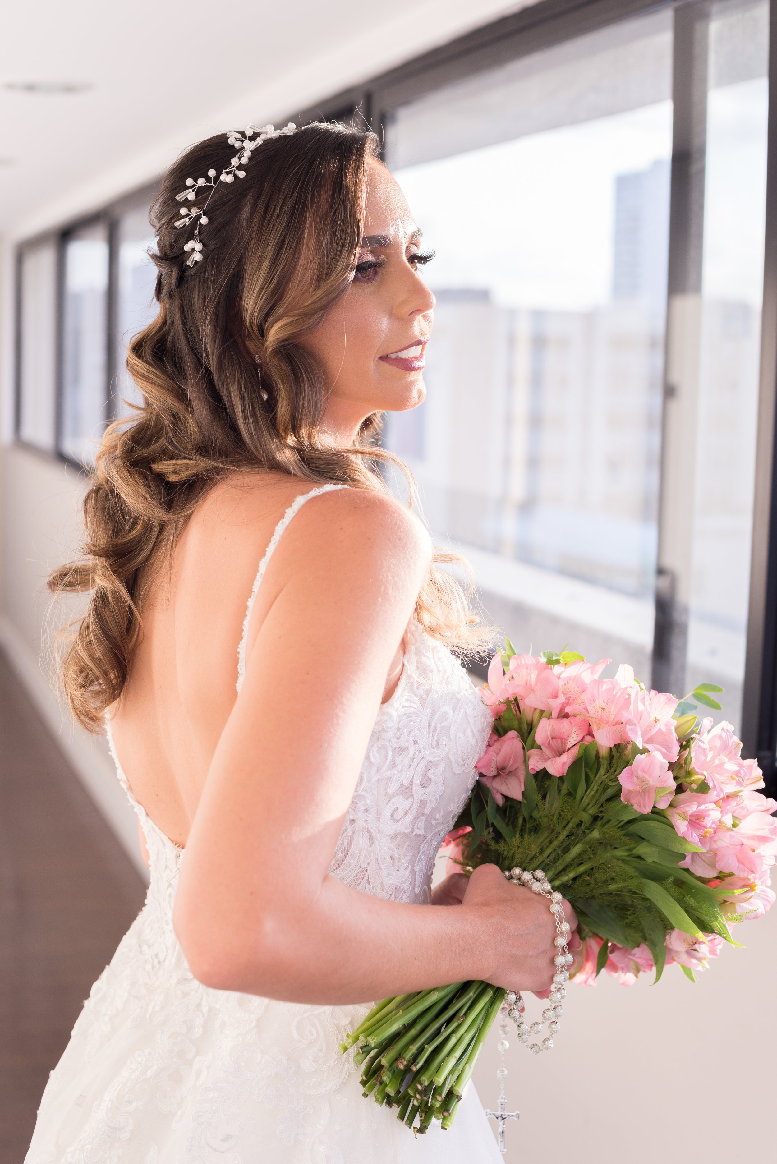 A bride in a white lace wedding dress holding a bouquet of pink flowers, standing by a large window.