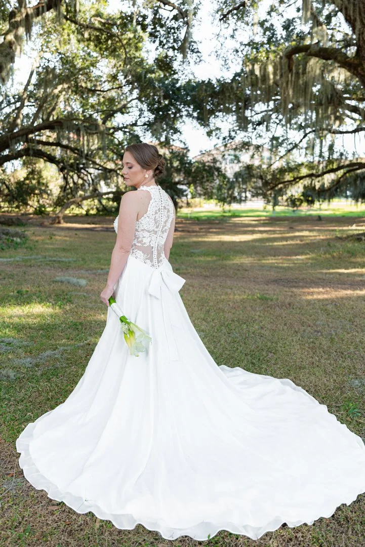 Bride in a white wedding dress holding calla lilies, standing outdoors under large trees with sunlight filtering through.