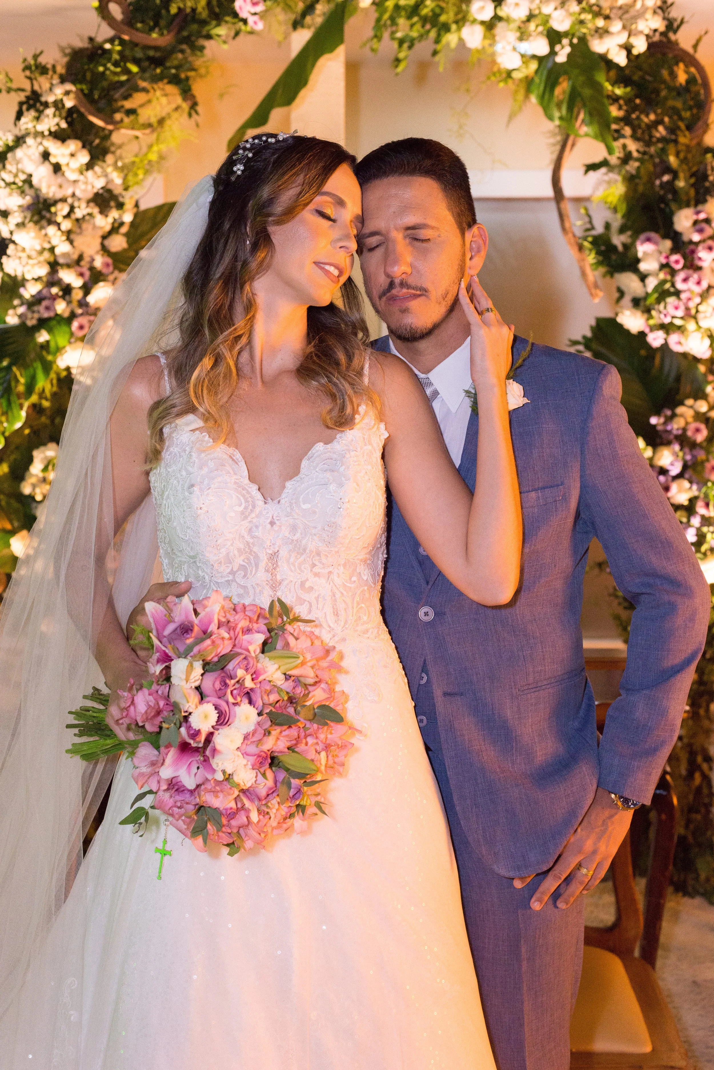 Bride in a white lace wedding gown holding a bouquet of pink and purple flowers, standing close to groom in a blue suit, both with eyes closed in a tender moment, surrounded by floral decorations.