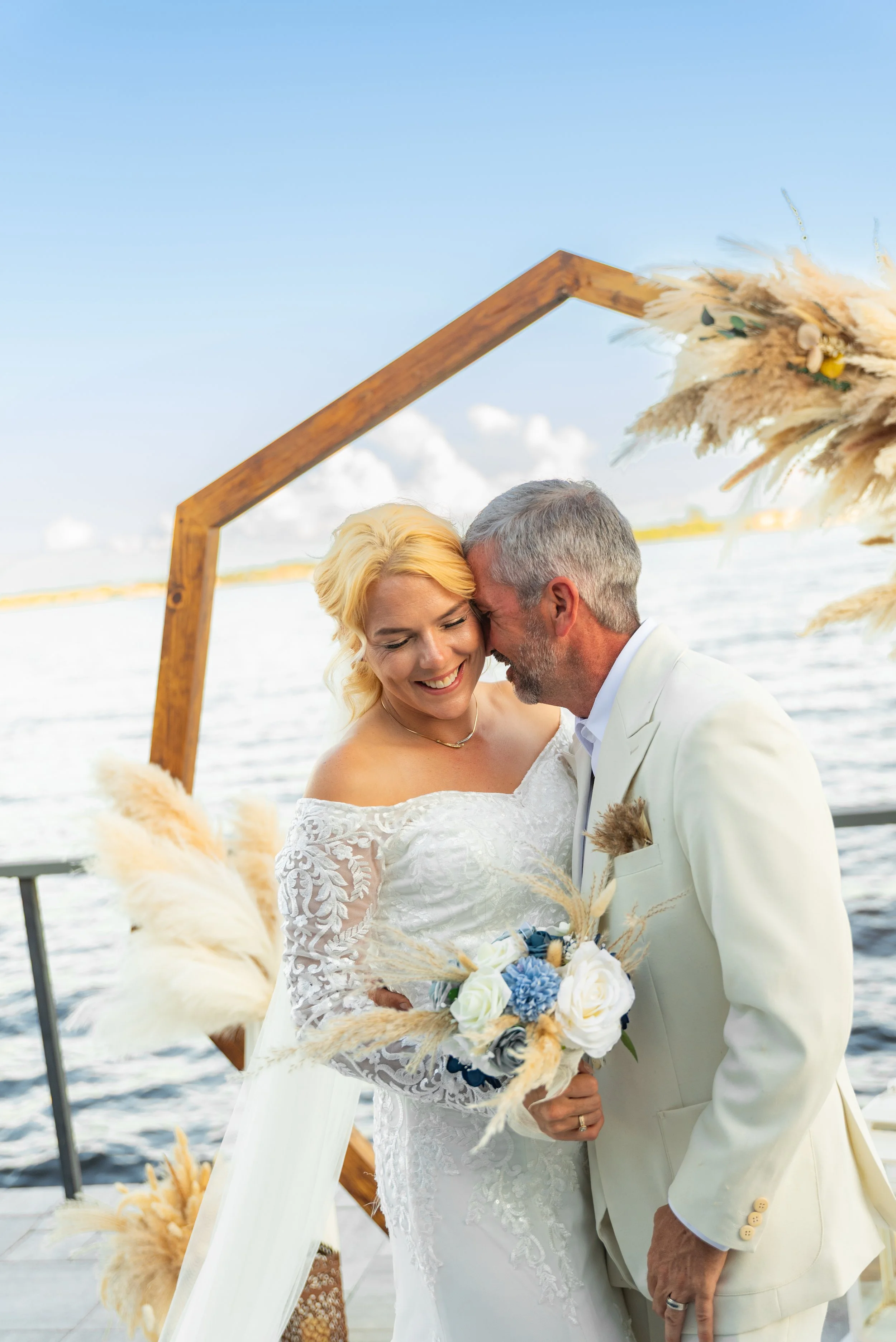 A couple in wedding attire sharing an intimate moment on a boat, with a decorative wooden arch and water in the background.