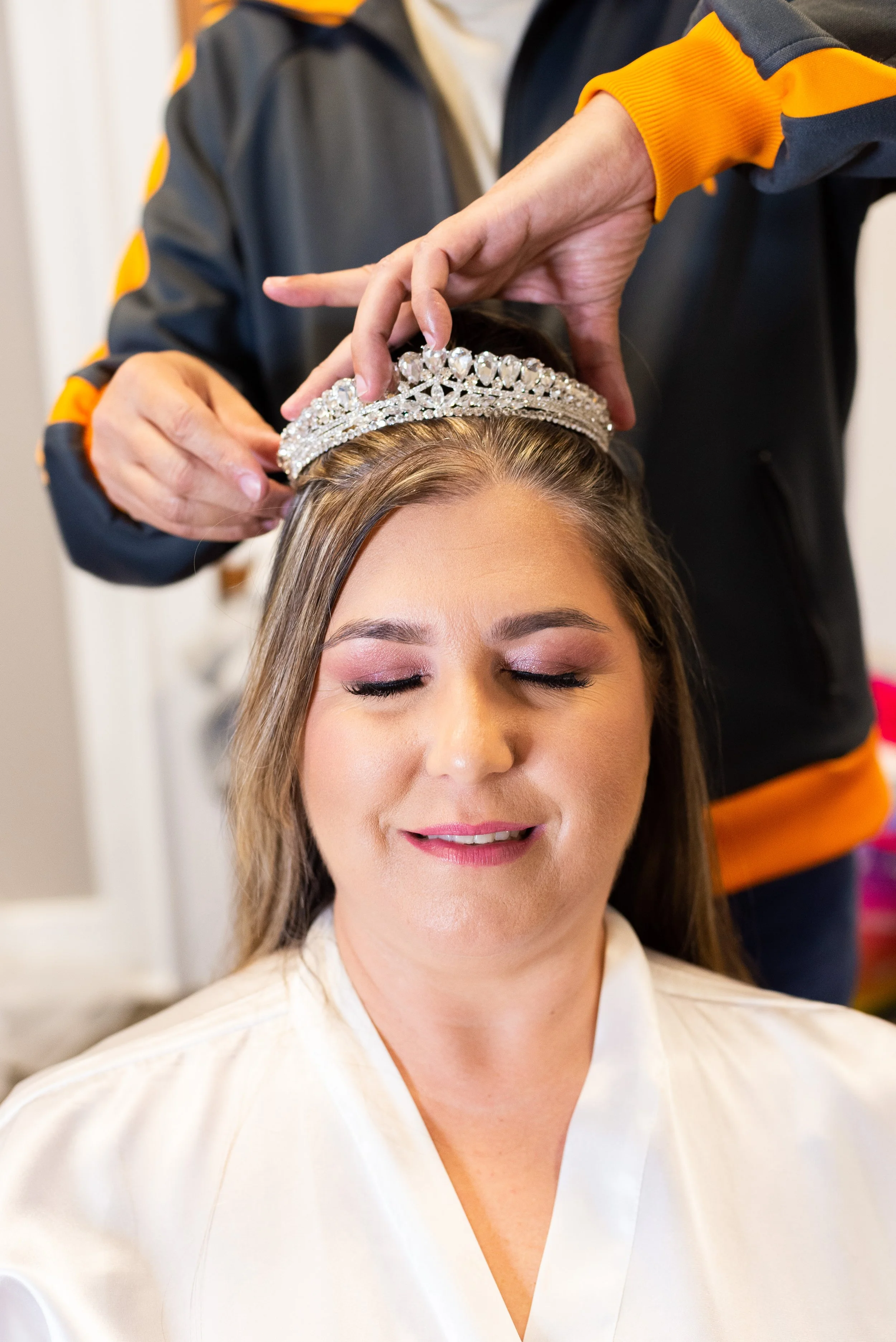 A woman with closed eyes wearing a tiara being adjusted by another person.