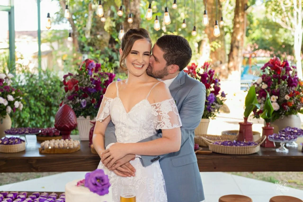 A bride and groom smiling and embracing at their wedding reception, with flowers and decorations in the background.