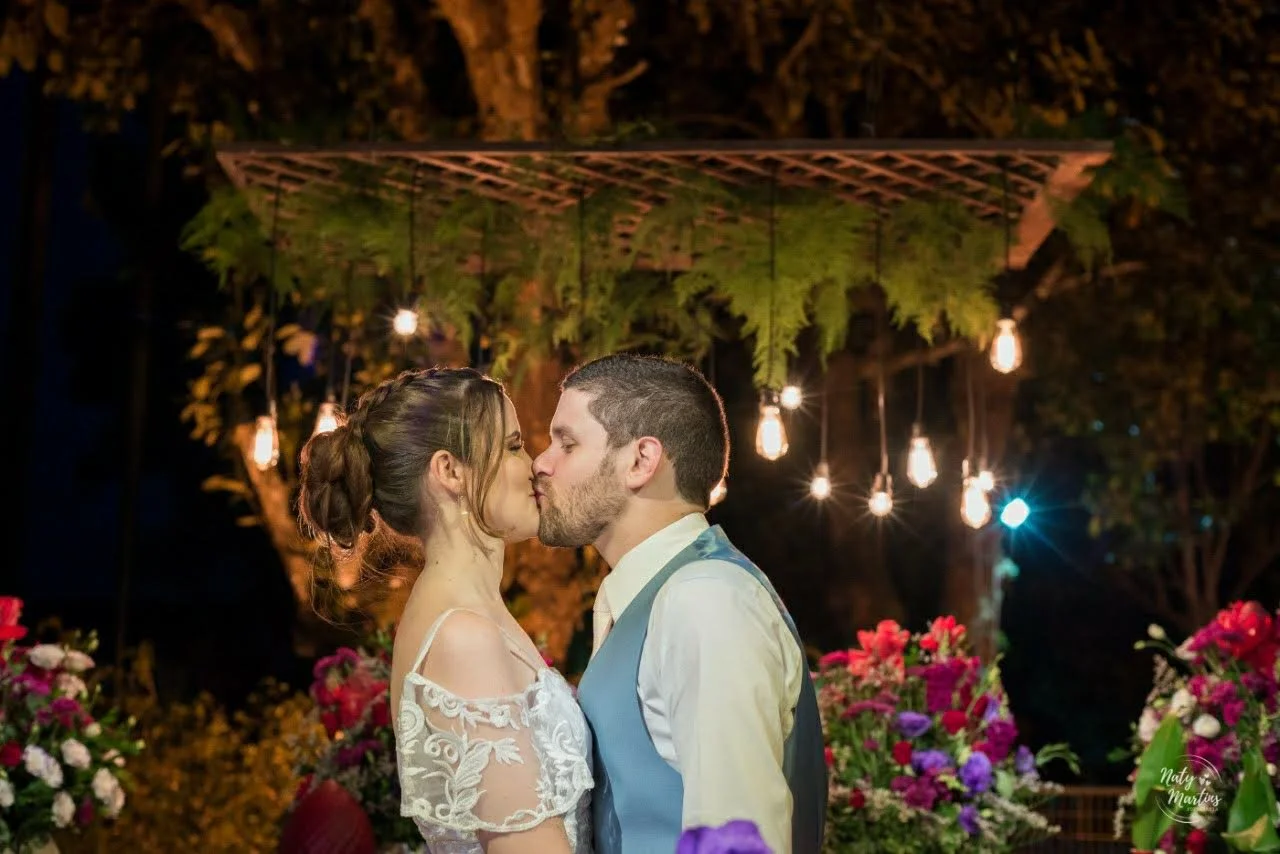 A bride and groom kissing at their wedding reception outdoors at night, with hanging lights and floral decorations in the background.