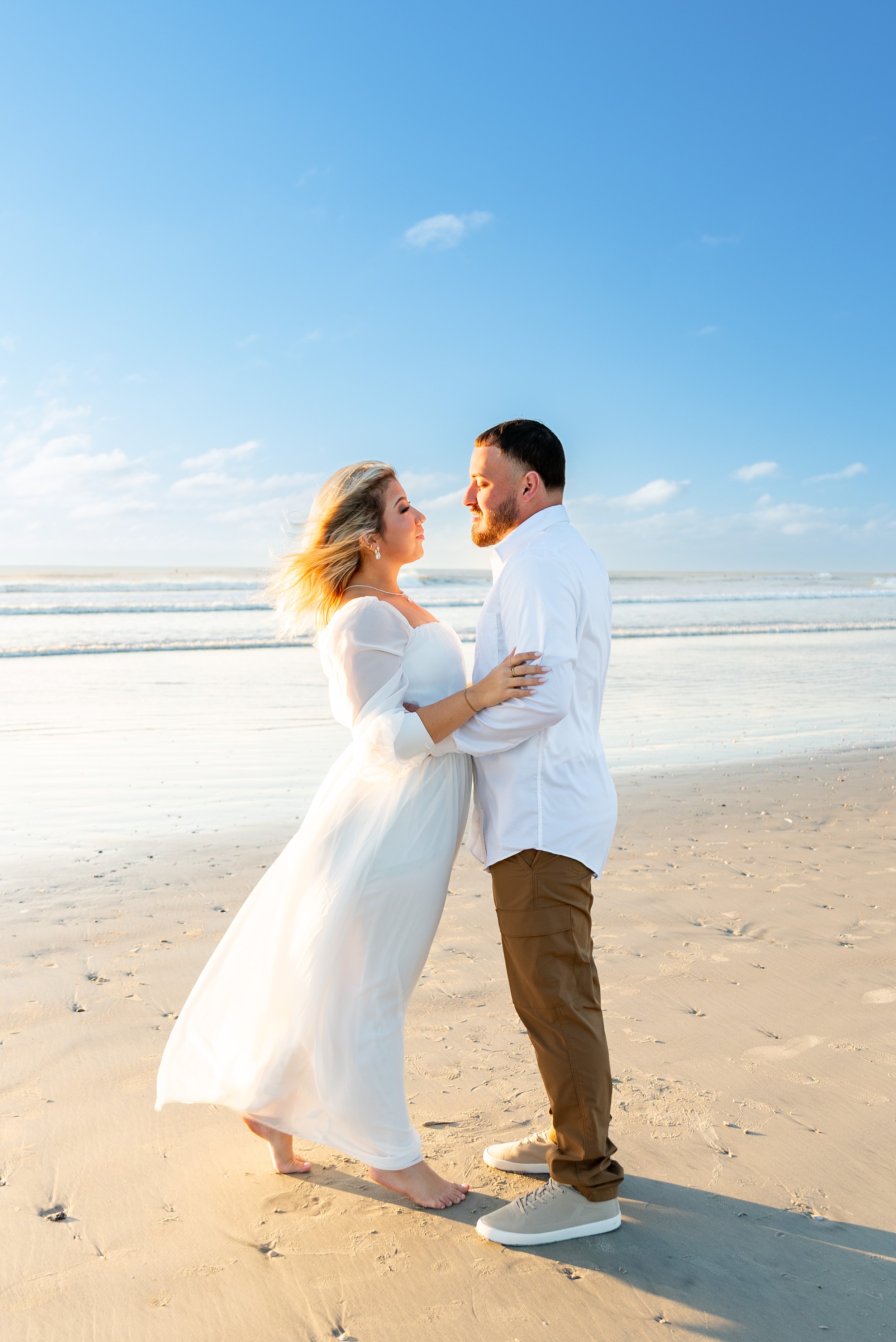 A couple standing close on a sandy beach, facing each other with their arms around each other, facing the ocean with a clear sky.