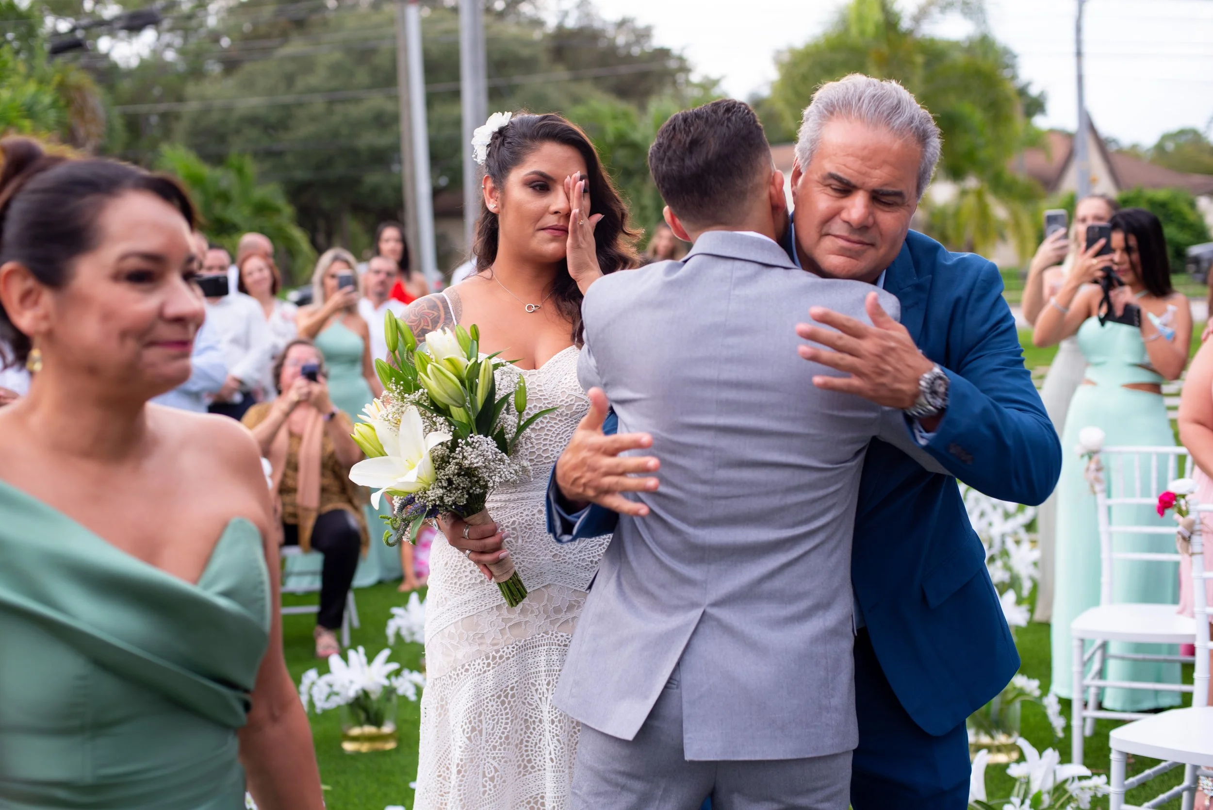 An emotional moment during a wedding ceremony with a man and a boy hugging, a woman with a bouquet of flowers wiping tears, and wedding guests in the background capturing the moment.