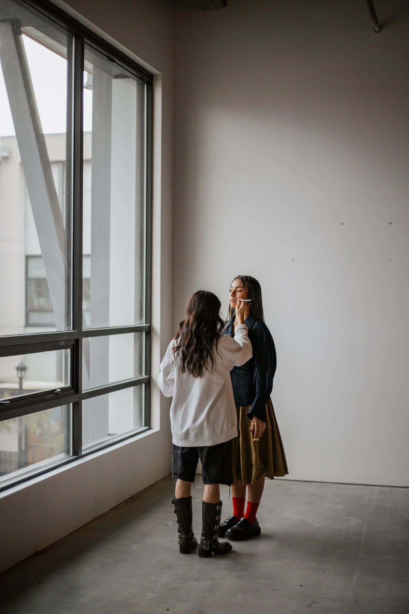 Person applying makeup to another person near a window in a minimalist room.