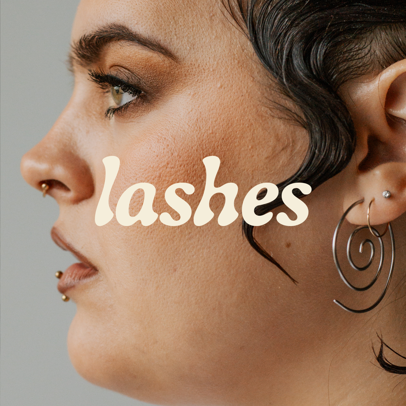 Close-up profile of a person with bold makeup, pierced nose, spiral earrings, and the word 'lashes' overlaid.