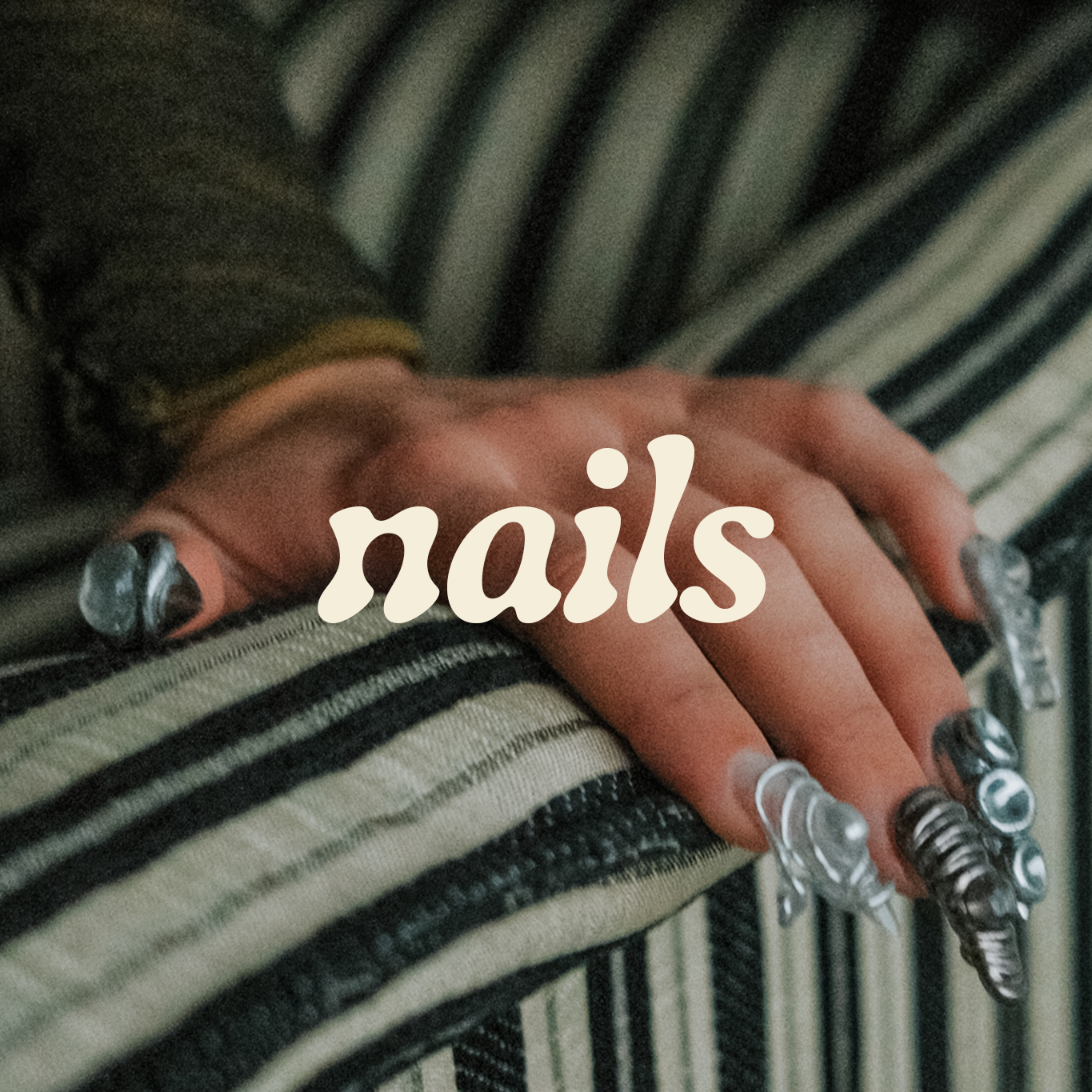 Hand with artistic nail designs on striped fabric, text "nails" overlay.