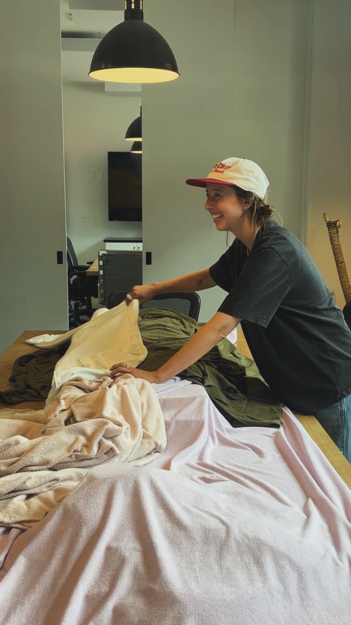 A woman with a white and red baseball cap and a dark t-shirt is making a bed with blankets on a wooden table, smiling.