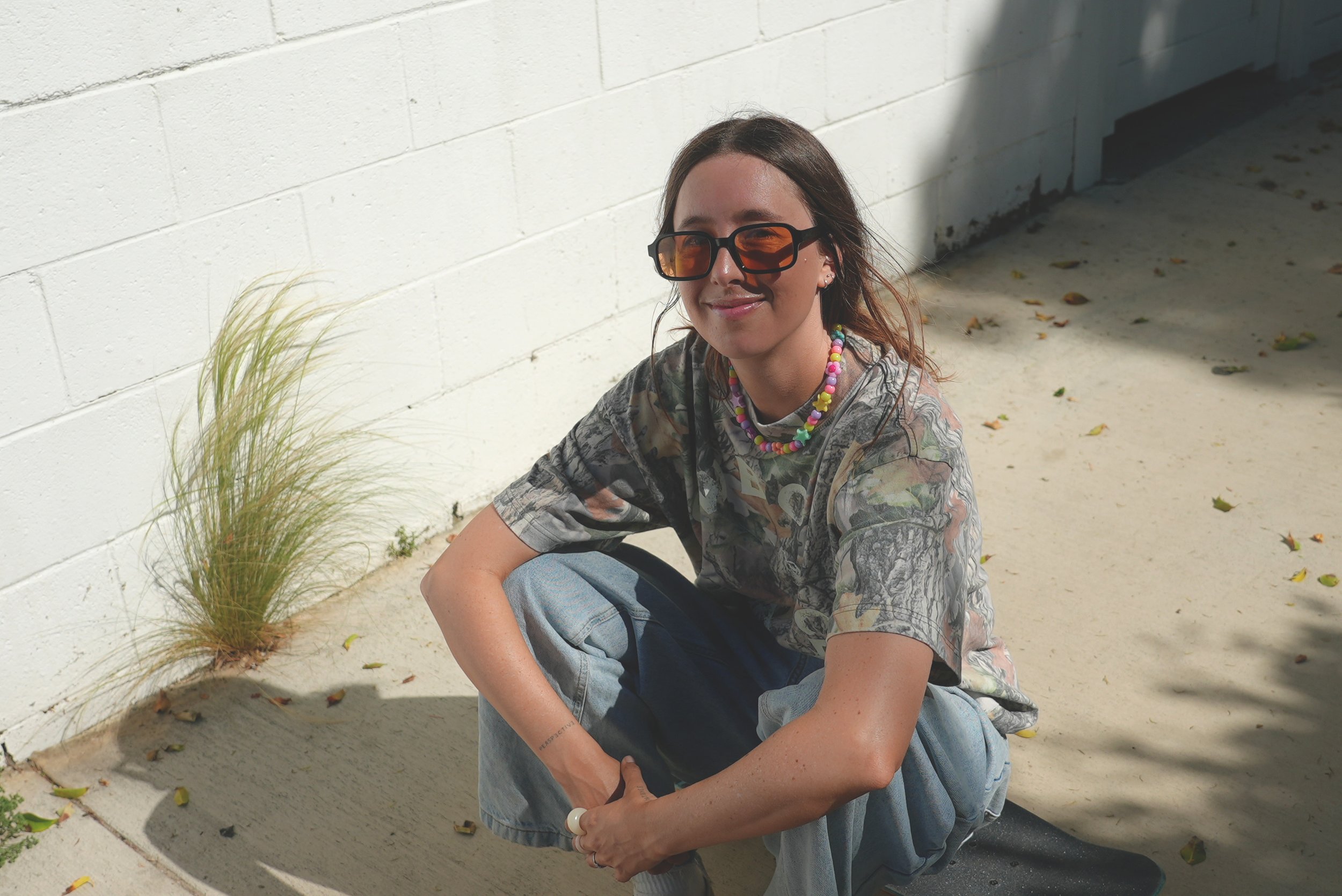A young woman with long brown hair, wearing orange-tinted sunglasses, a colorful beaded necklace, a patterned T-shirt, and light blue jeans, crouching outside on sand with a white brick wall behind her.