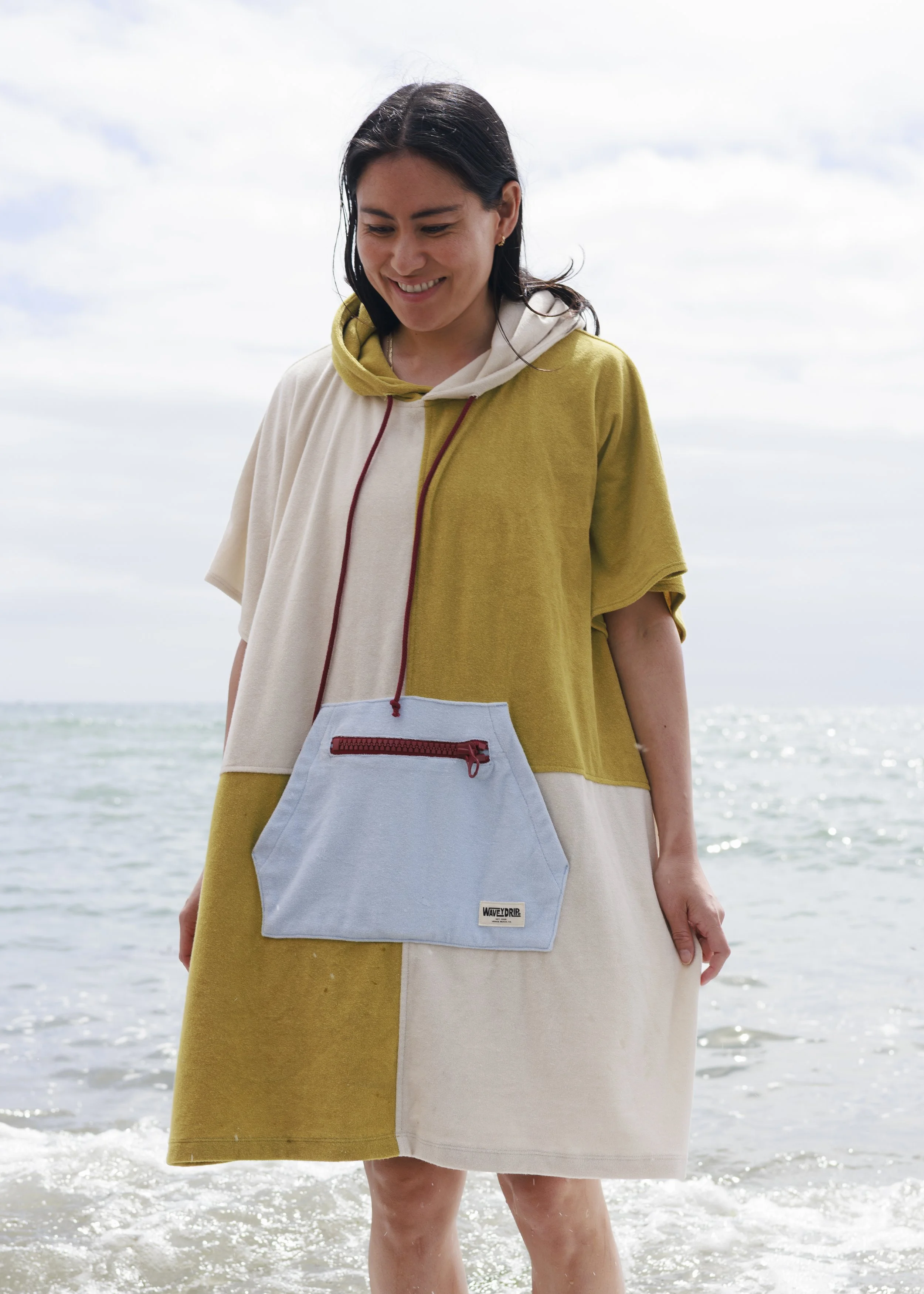 Woman smiling at the beach, wearing a color-block hoodie dress with a large light blue pouch attached around her neck, standing in shallow water with the ocean in the background.