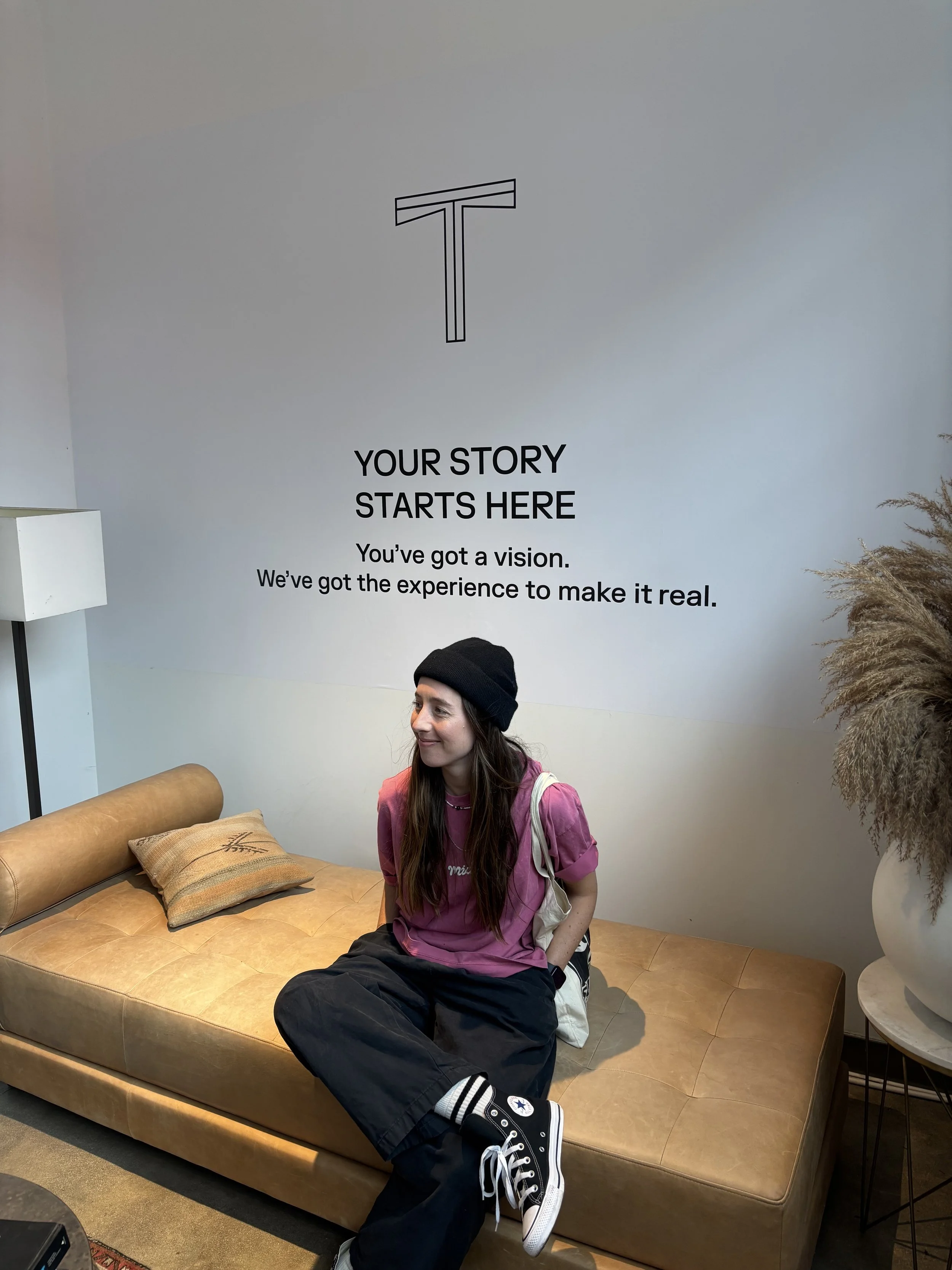 A young woman in casual clothing, including a black beanie, pink t-shirt, black pants, and black Converse sneakers, is sitting on a tan leather couch with a beige cushion. She is in a modern, minimalist room with a white wall that has motivational text and a simple line art logo at the top. There is a white standing lamp to the left and a large white vase with dried pampas grass to the right.