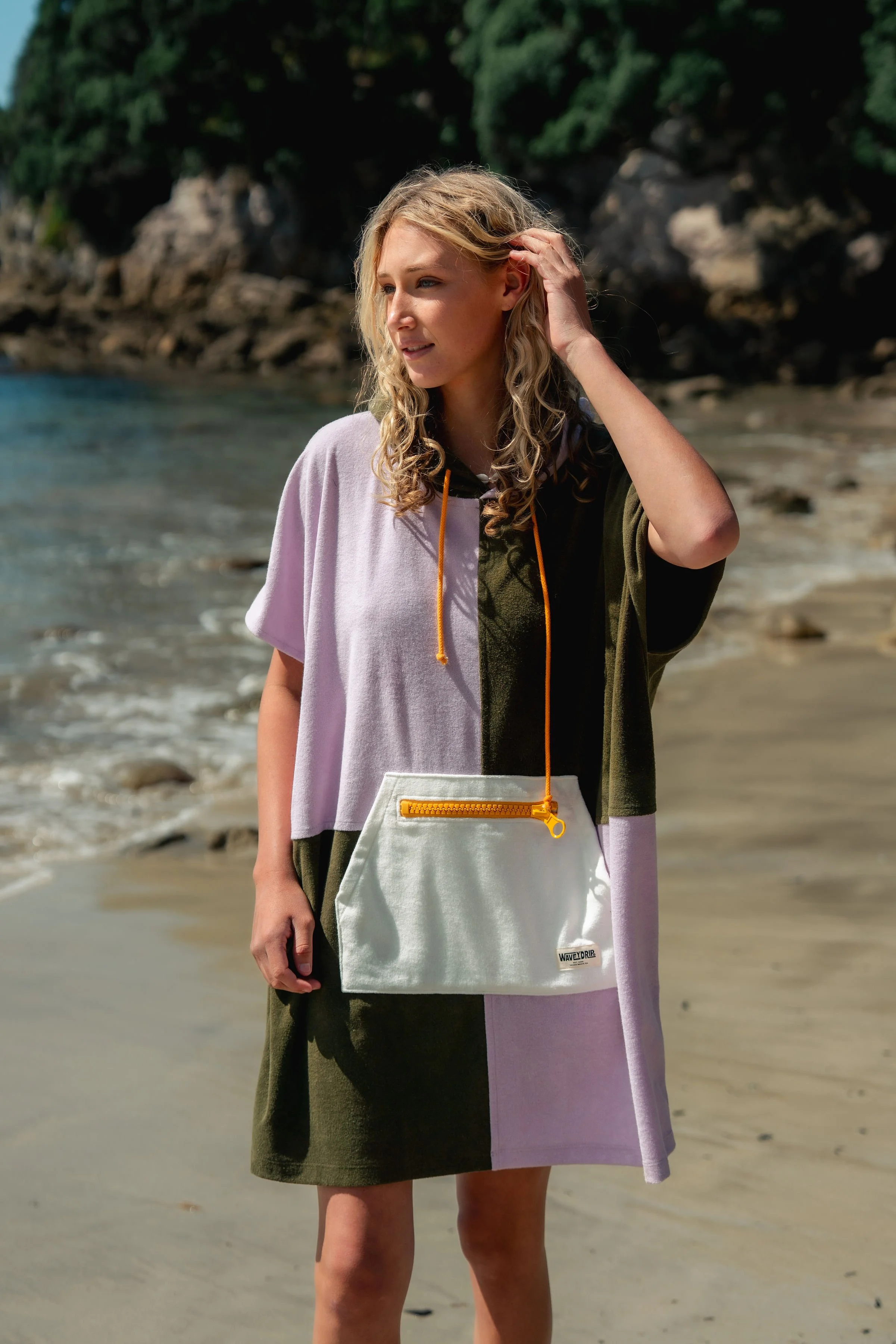 A young woman with curly blonde hair standing on a beach with rocky shoreline and trees in the background, wearing a color-blocked batik towel dress and holding a white bag with an orange zipper and strap.