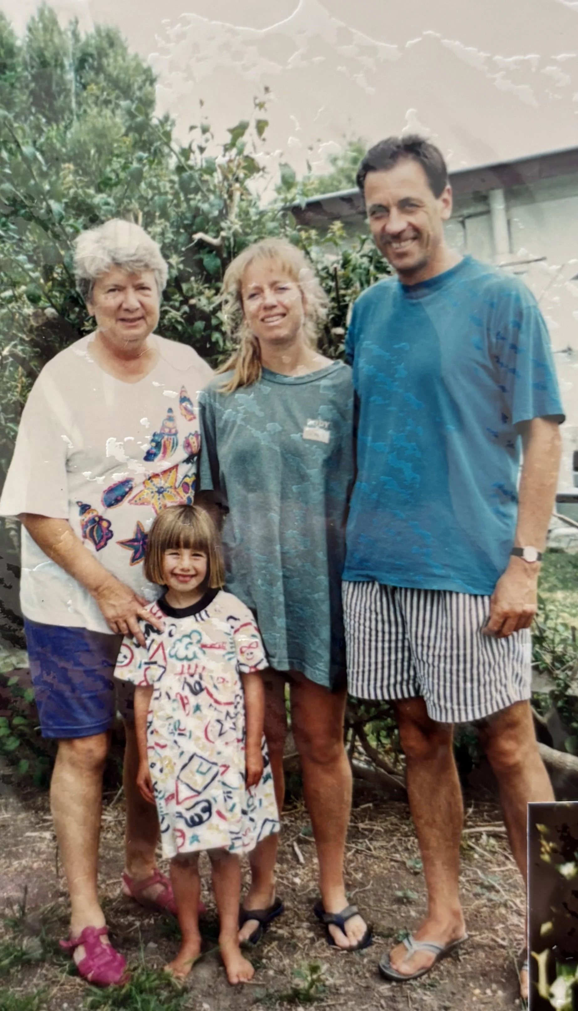 A family of five standing outdoors in a garden with trees and a building in the background. The group includes an elderly woman, a middle-aged woman, a middle-aged man, and a young girl, all smiling.