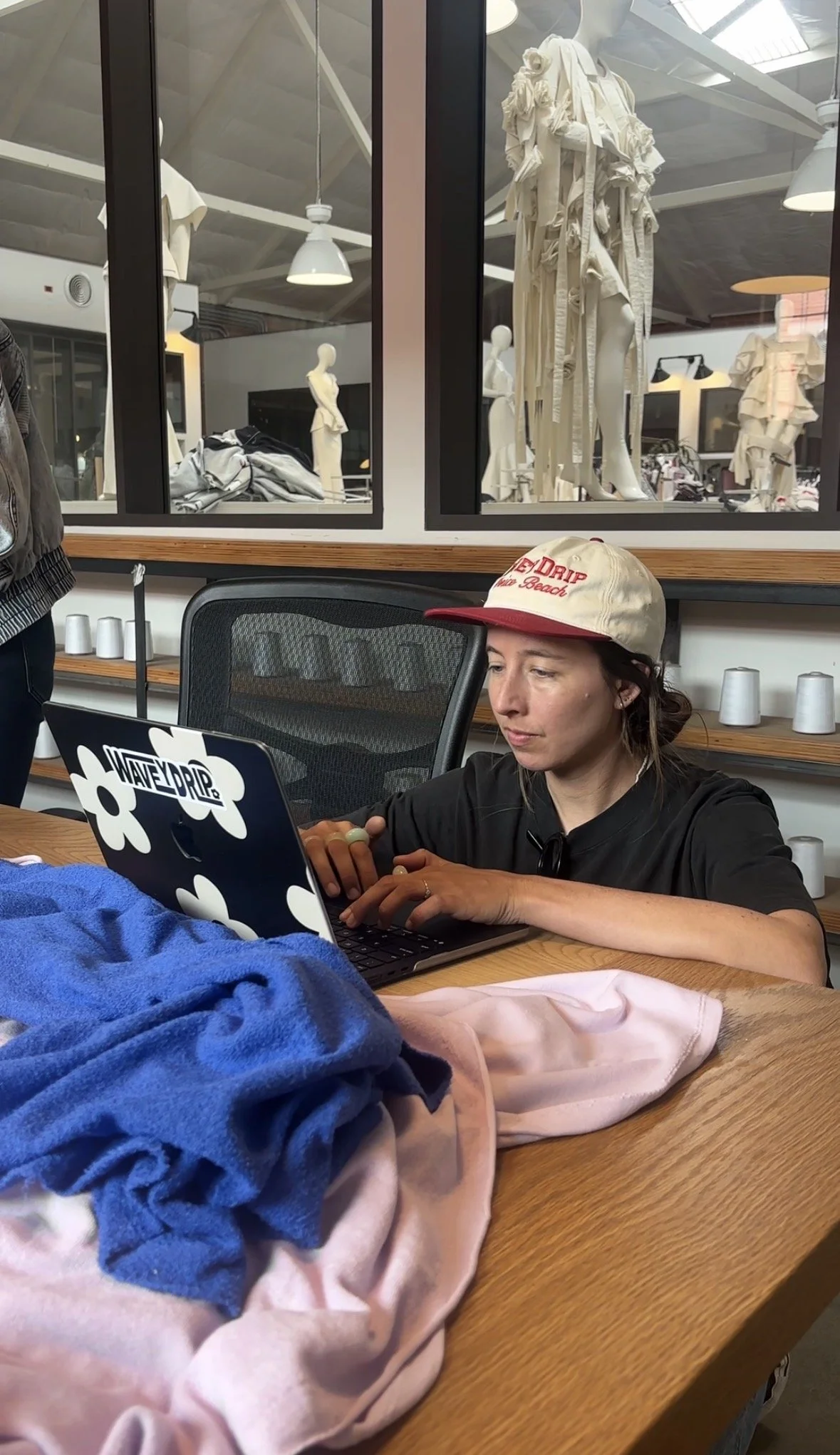 A woman wearing a white cap with red text, sitting at a desk with a laptop, with clothing and fabrics in front of her.