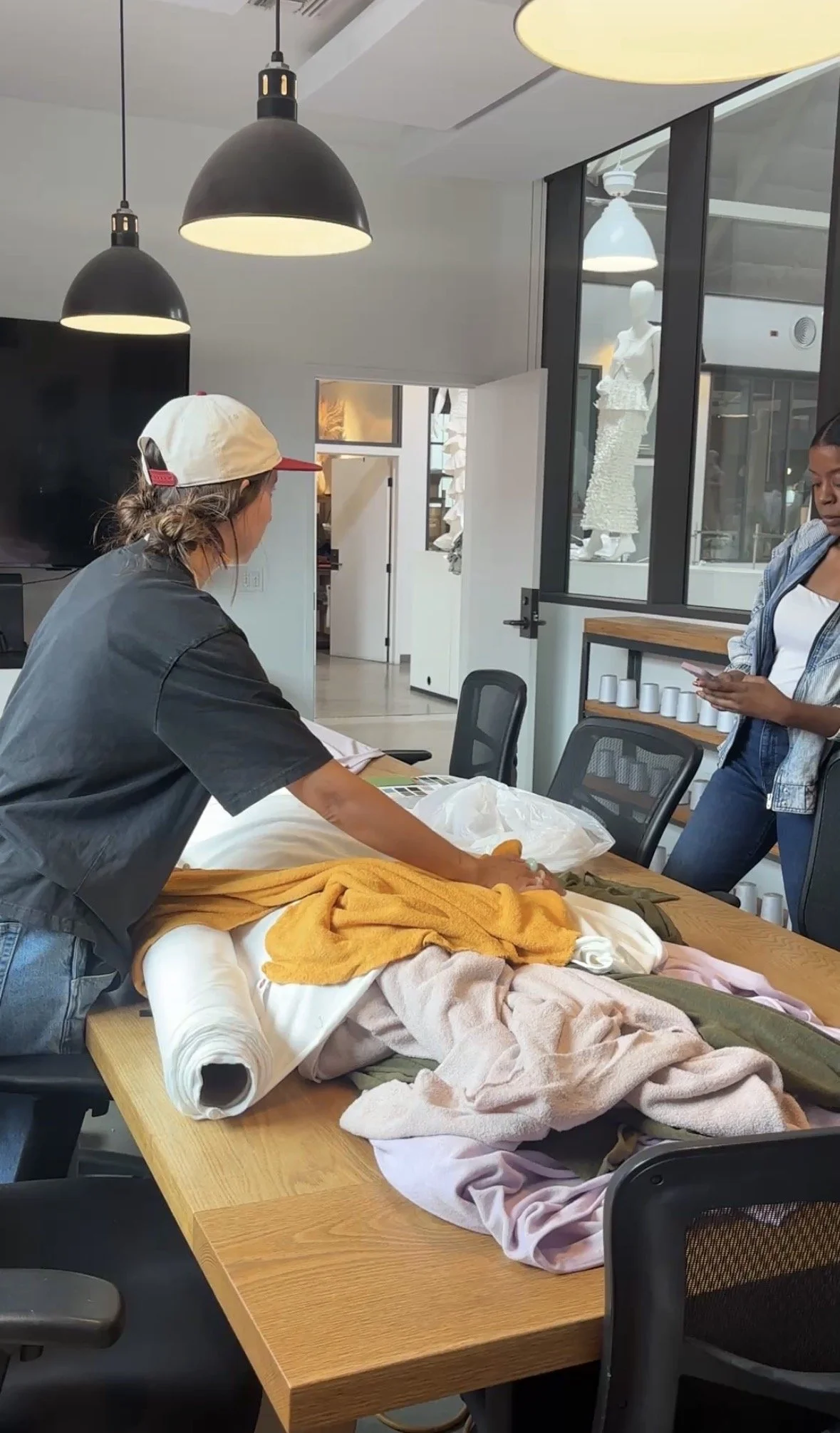 Person arranging clothing and textiles on a table in an office or studio space while another person looks on.