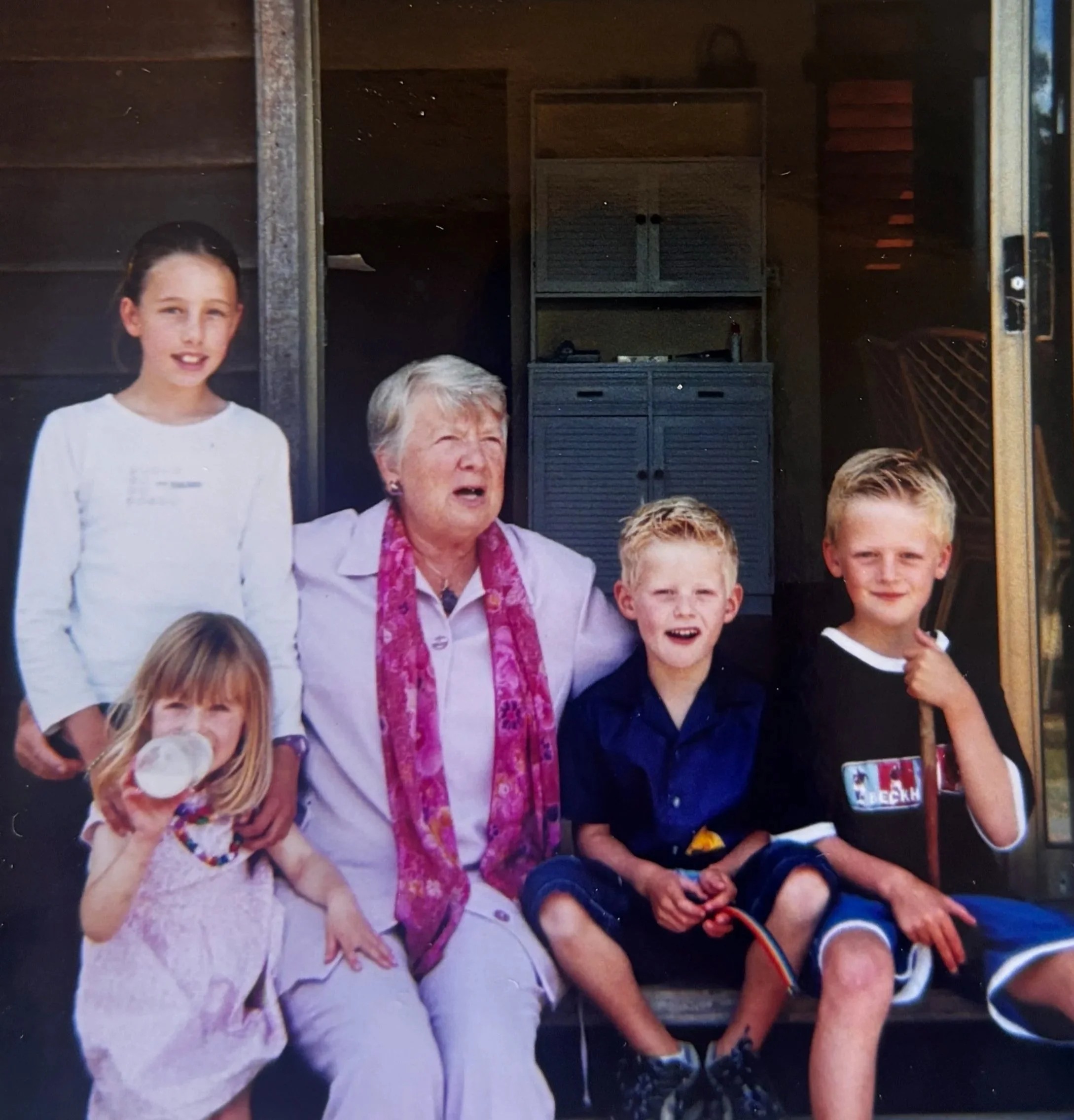 A group of four children and an elderly woman sitting on a porch. One girl is standing in a white long-sleeve shirt, another girl is sitting and drinking a beverage, and two boys are sitting, one holding a stick. The elderly woman is wearing a light purple outfit and a pink scarf, and appears to be mid-sentence or singing.