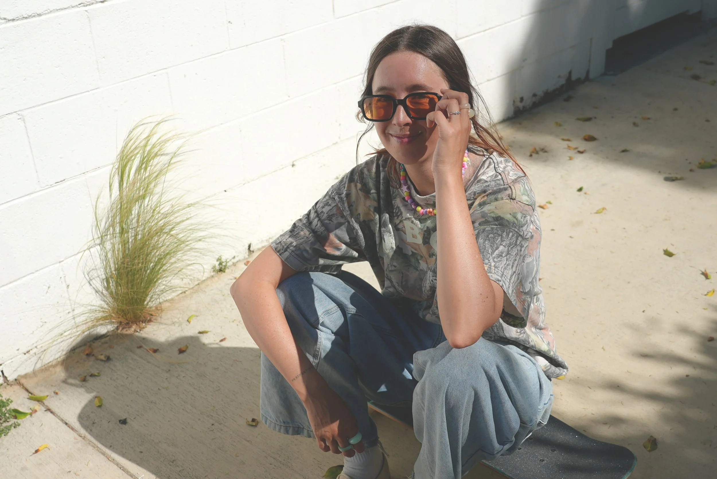 A young woman with long brown hair squatting outside on a sunny day, wearing orange-tinted sunglasses, a colorful beaded necklace, a printed t-shirt, baggy jeans, and white sneakers.