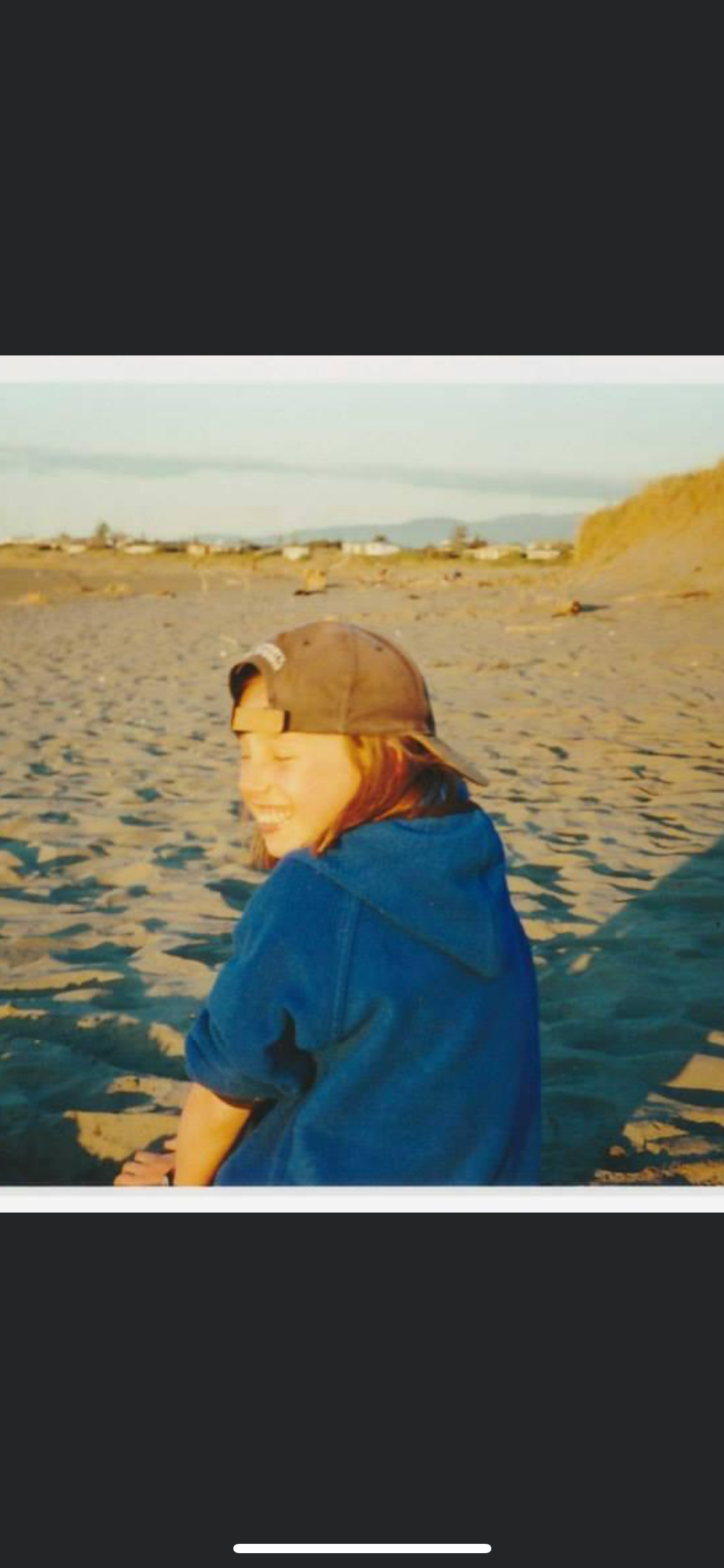 Person sitting on sandy beach, smiling, wearing a dark baseball cap and a blue hoodie, with distant buildings and mountains in the background during sunset.
