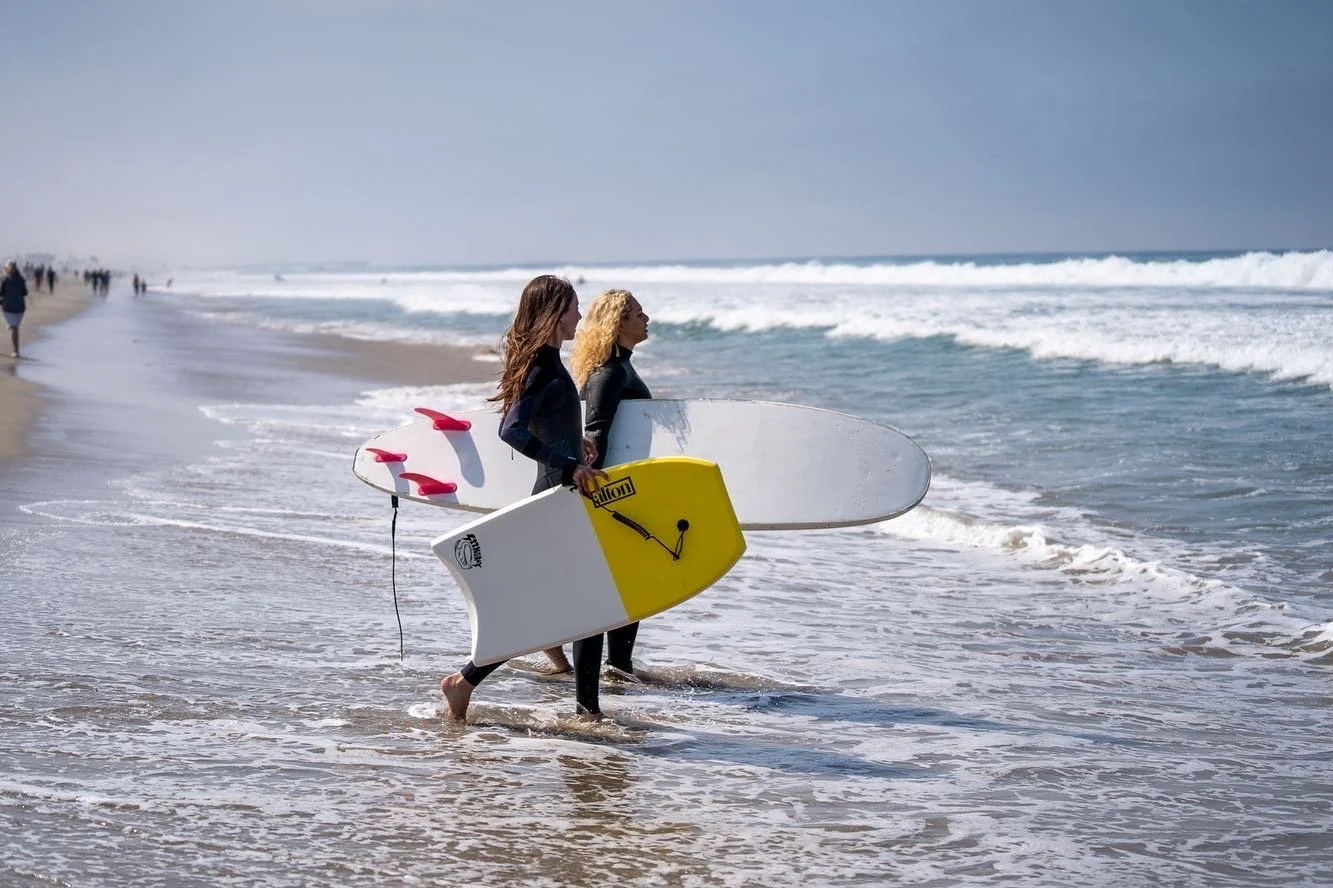 Two women with surfboards walking into the ocean on a sandy beach, with people in the distance and waves in the background.
