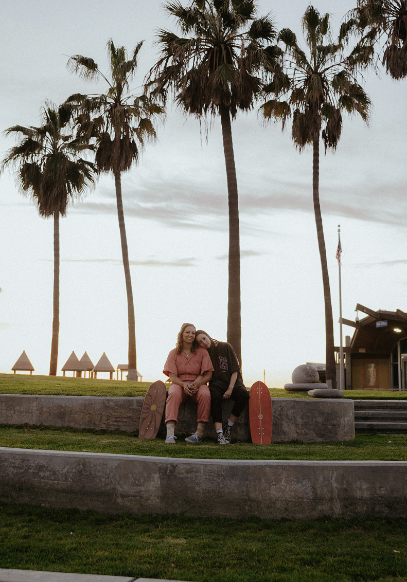 Two people sitting on a low concrete wall in front of tall palm trees during sunset or dusk, with a cloudy sky and a flagpole with an American flag in the background.