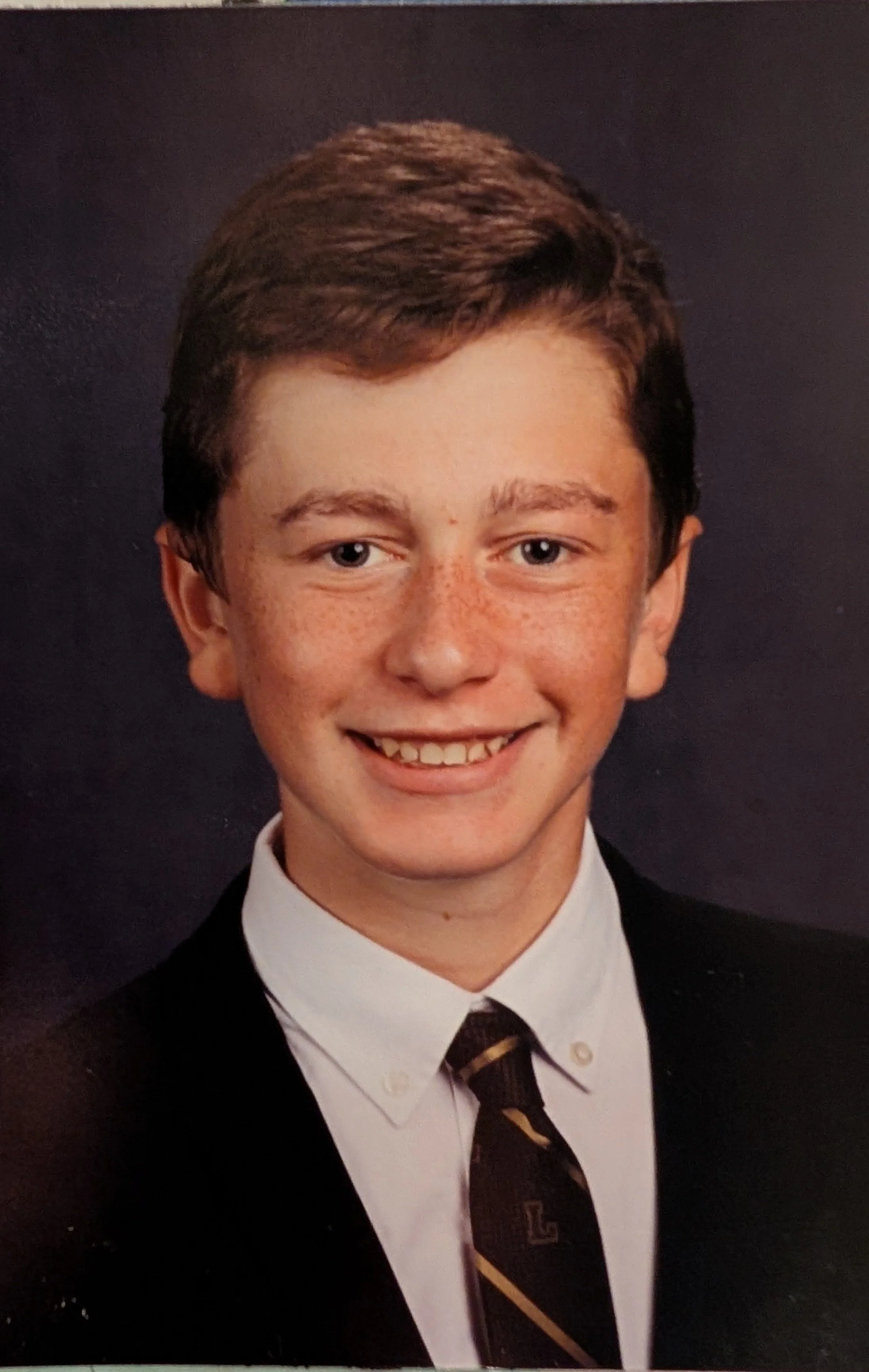 A young boy dressed in a school uniform, including a black blazer, white shirt, and a black and gold striped tie, smiling at the camera against a dark background.