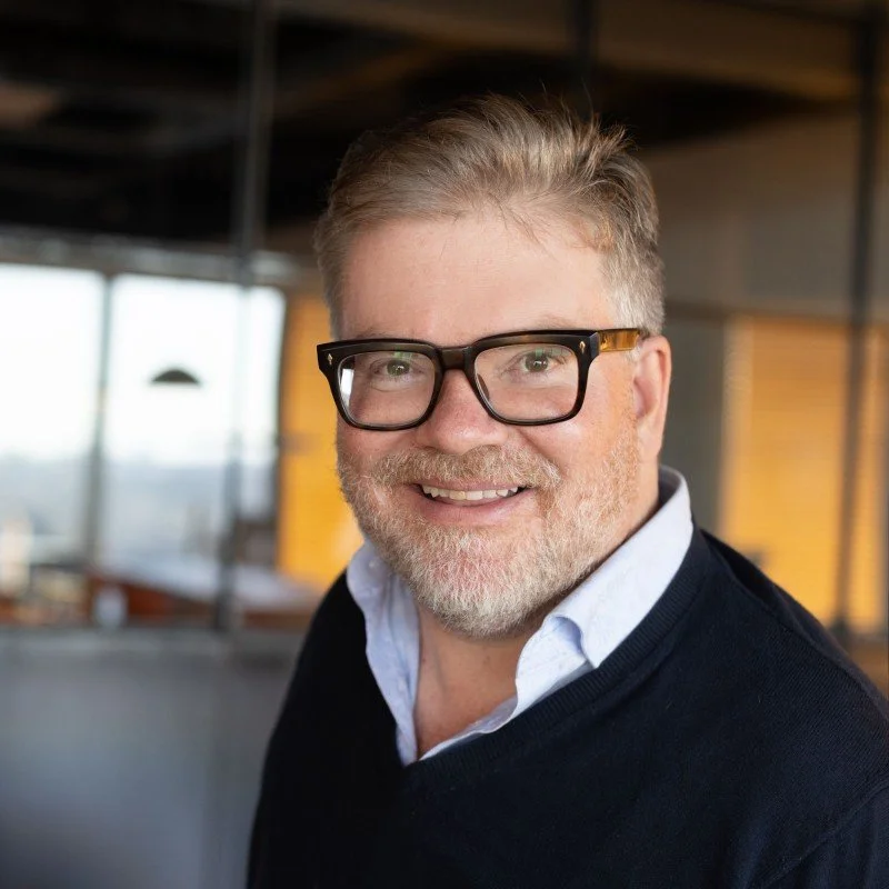 A smiling middle-aged man with glasses, short light brown hair, and a beard, wearing a white collared shirt and a dark sweater, in an indoor setting with large windows in the background.