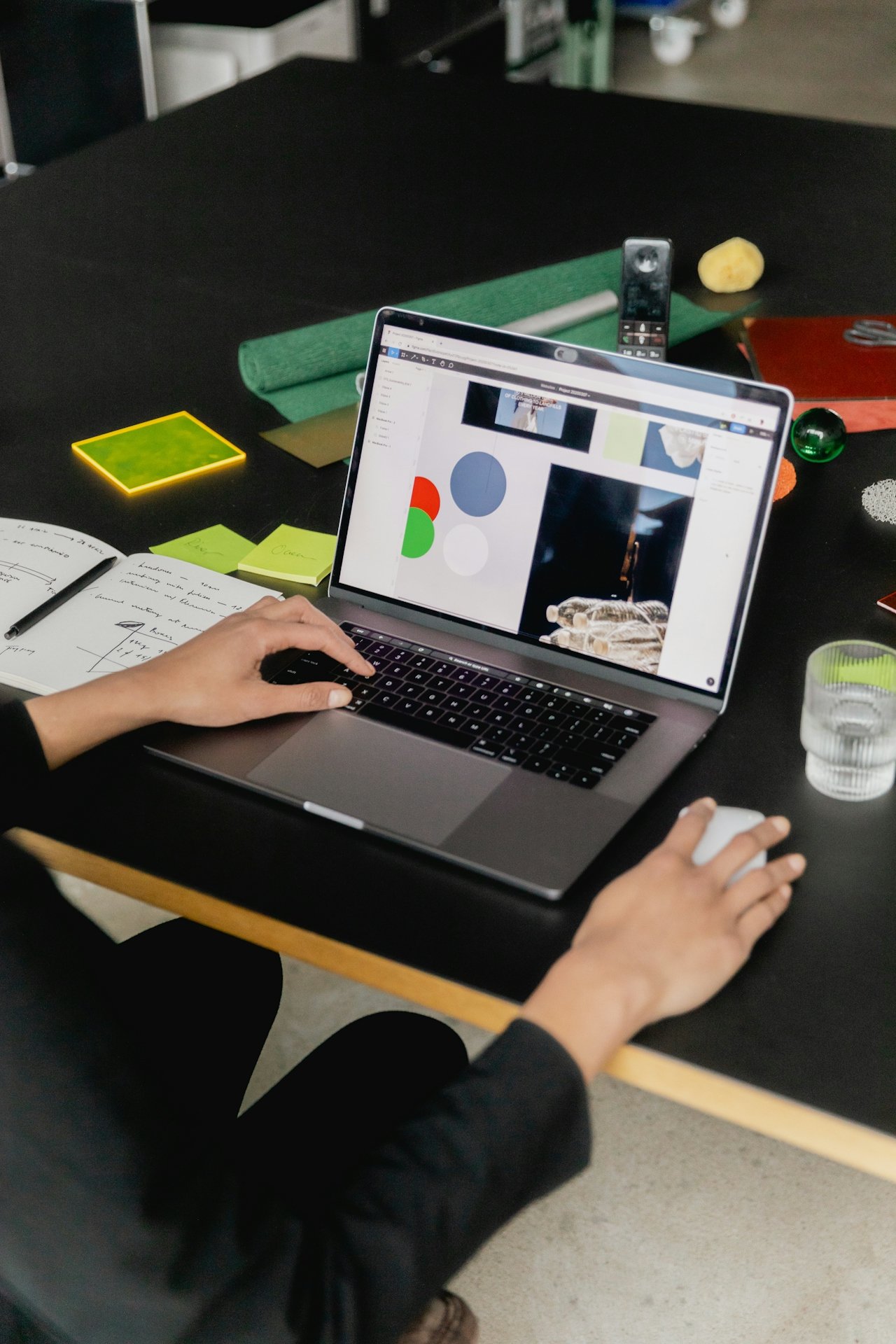 A person working on a laptop at a black table with various stationery items, sticky notes, a glass of water, and a remote control.