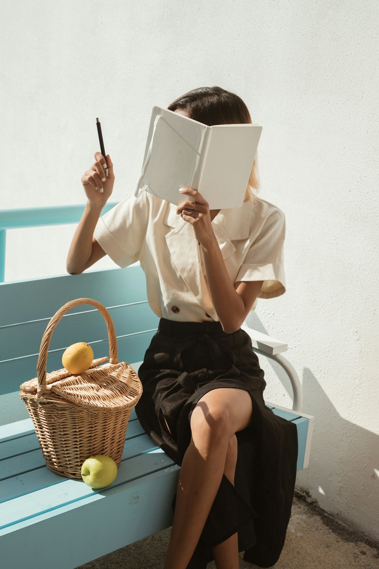 A woman sitting on a light blue bench outdoors, holding a notebook and pen, with two green apples and a wicker basket nearby.