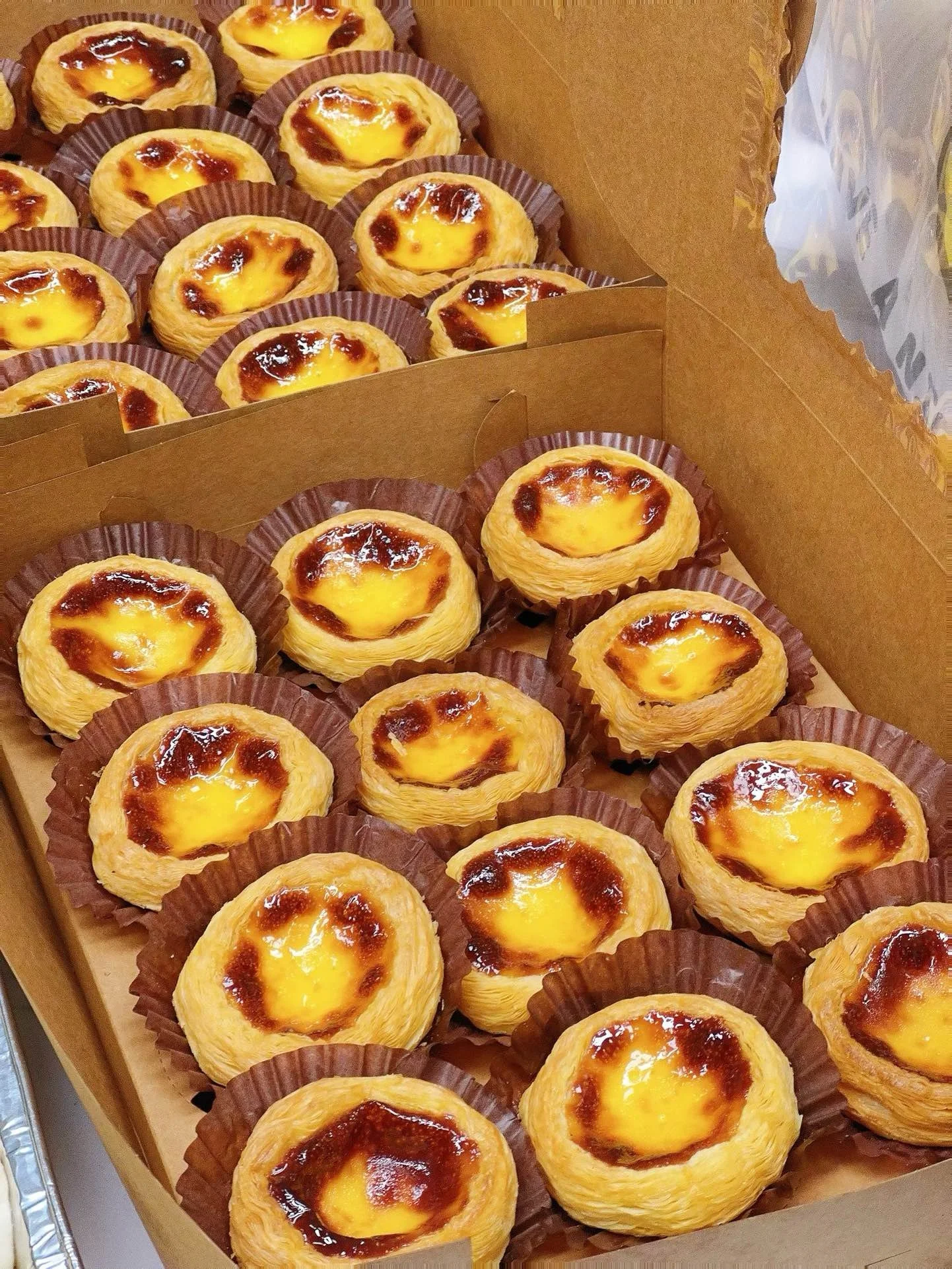 A box of freshly baked Portuguese egg tarts, each in a brown paper cup, with golden custard filling and caramelized tops.