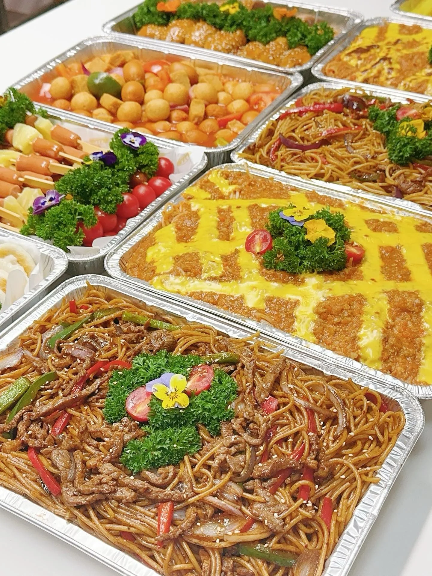 Assorted catering food dishes in aluminum trays, including fried rice, lo mein, sweet and sour chicken, and vegetable stir-fry garnished with parsley and edible flowers.