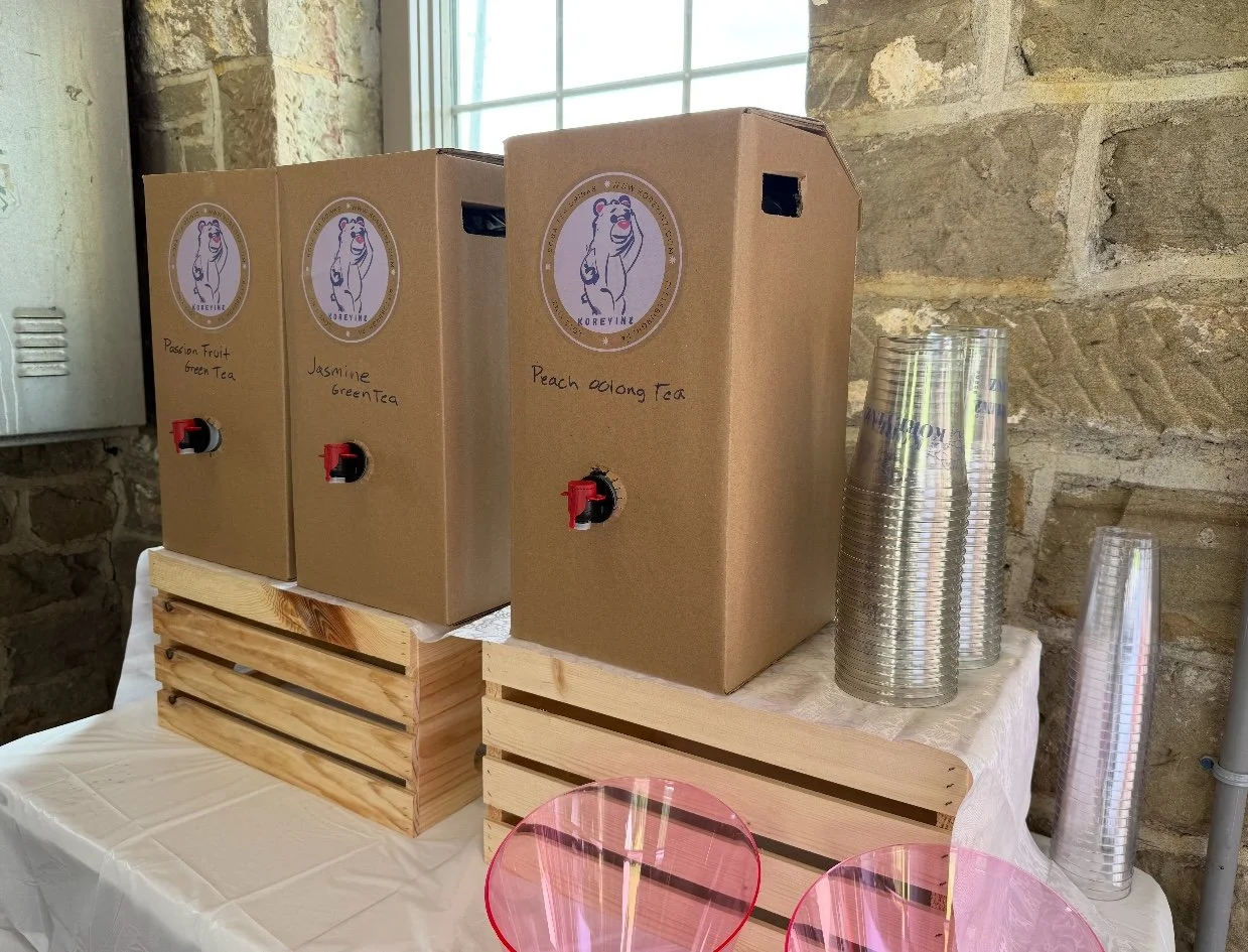 Three cardboard boxes labeled with flavored tea, passing fruit green tea, jasmine green tea, and peach oolong tea, are on a table with stacked clear plastic cups and pink and clear glasses. The table is covered with a white tablecloth and has wooden 