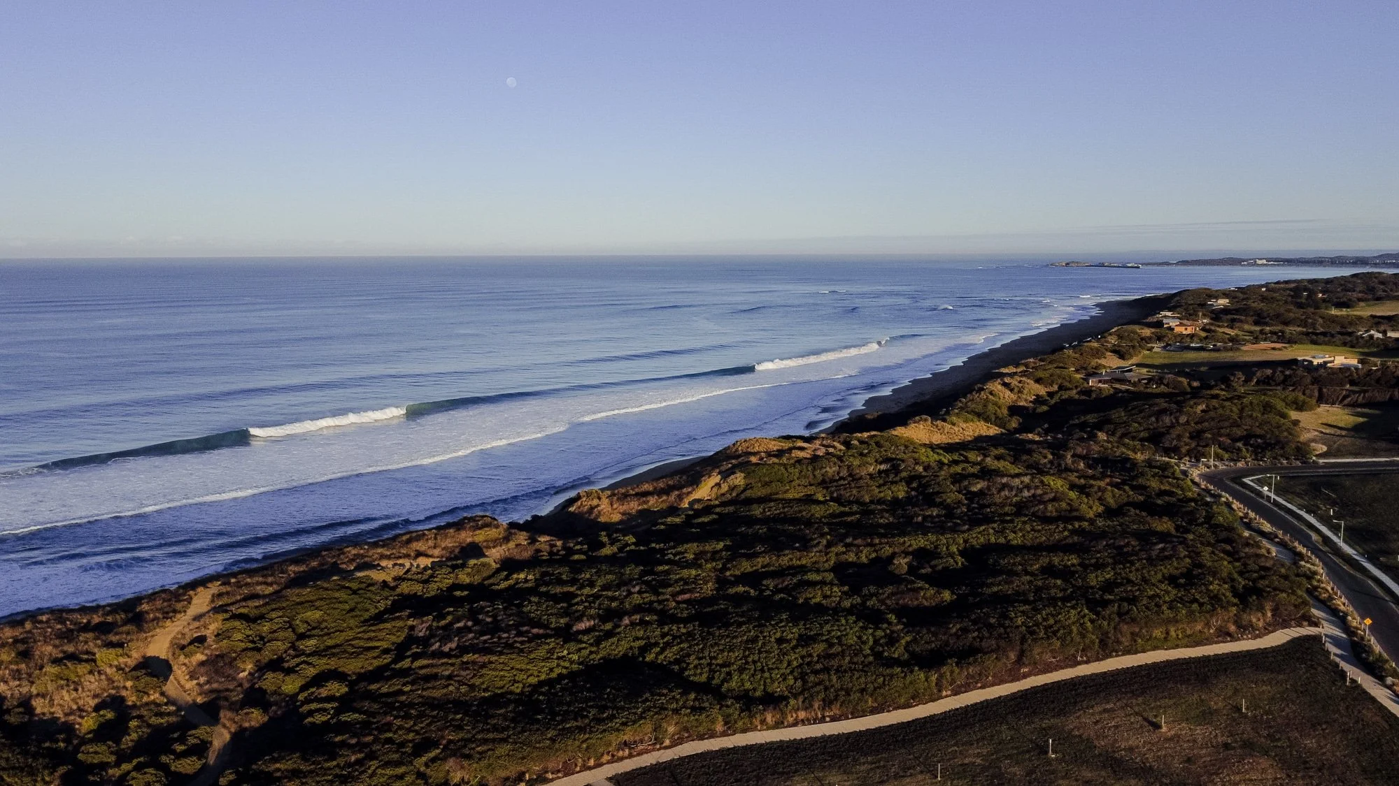 An aerial view of a coastline with ocean waves reaching the shore, lush green hills, and a winding road along the coast under a clear sky.