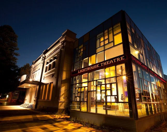 Nighttime exterior view of Lighthouse Theatre, a modern glass building with illuminated interior, adjacent to a classical building, and a tree on the left.