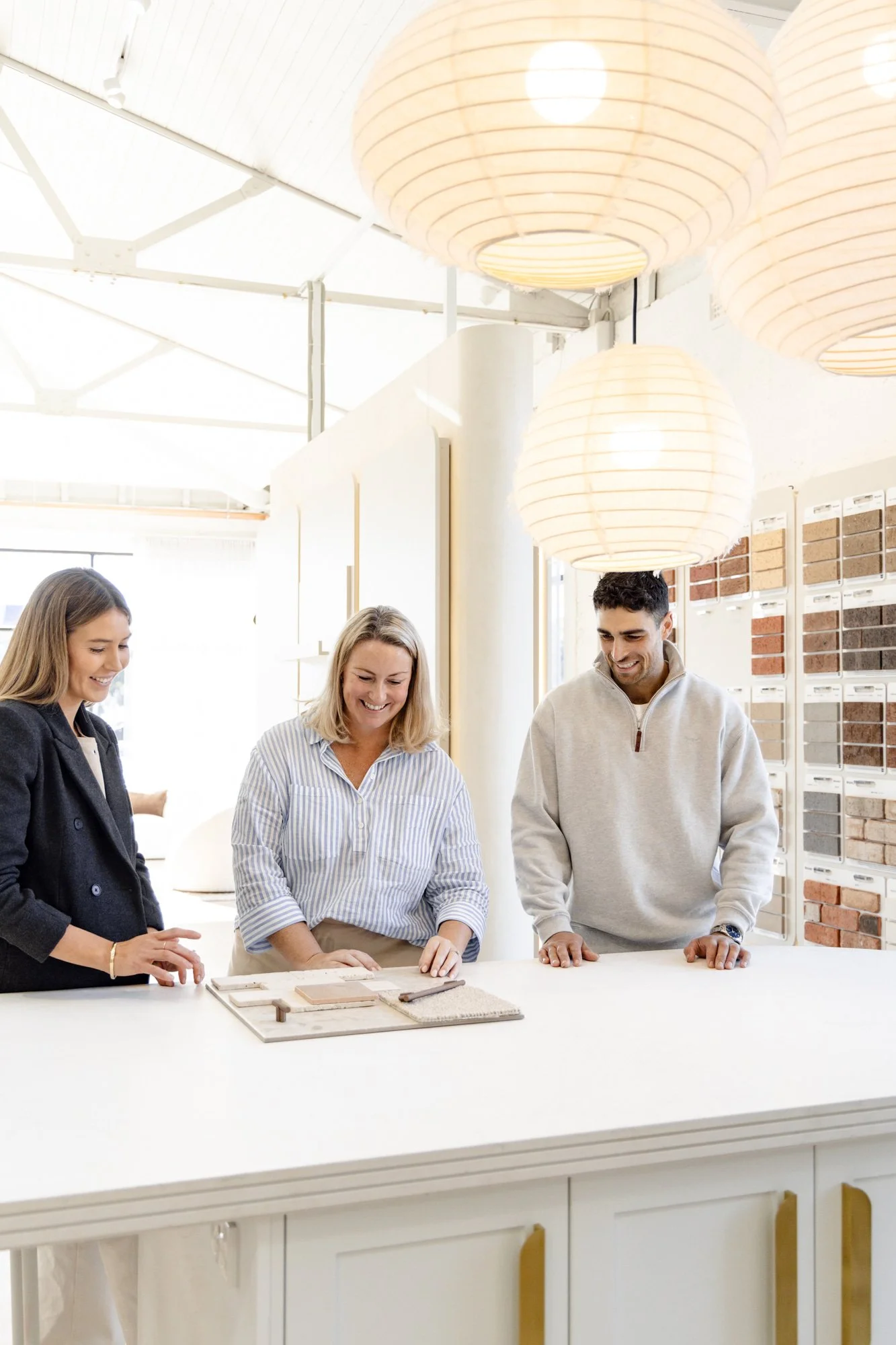 Three people smiling and looking at design samples on a table in a bright, modern interior with hanging lights and wall display of brick samples.