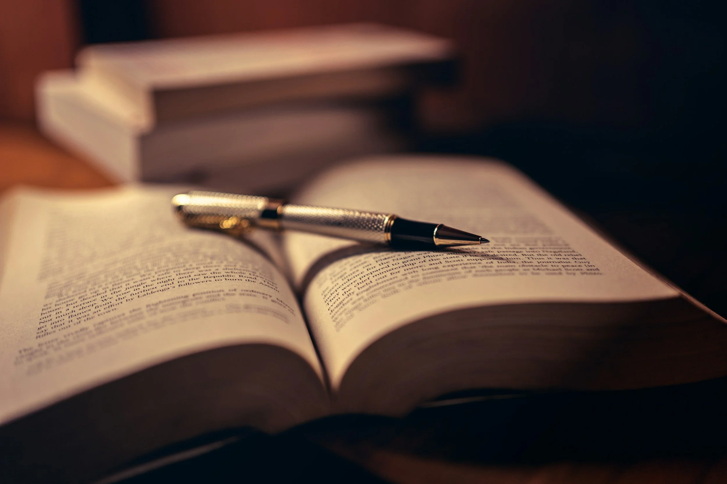 An open book with printed text, a metallic pen resting on the pages, and a blurry stack of books in the background.