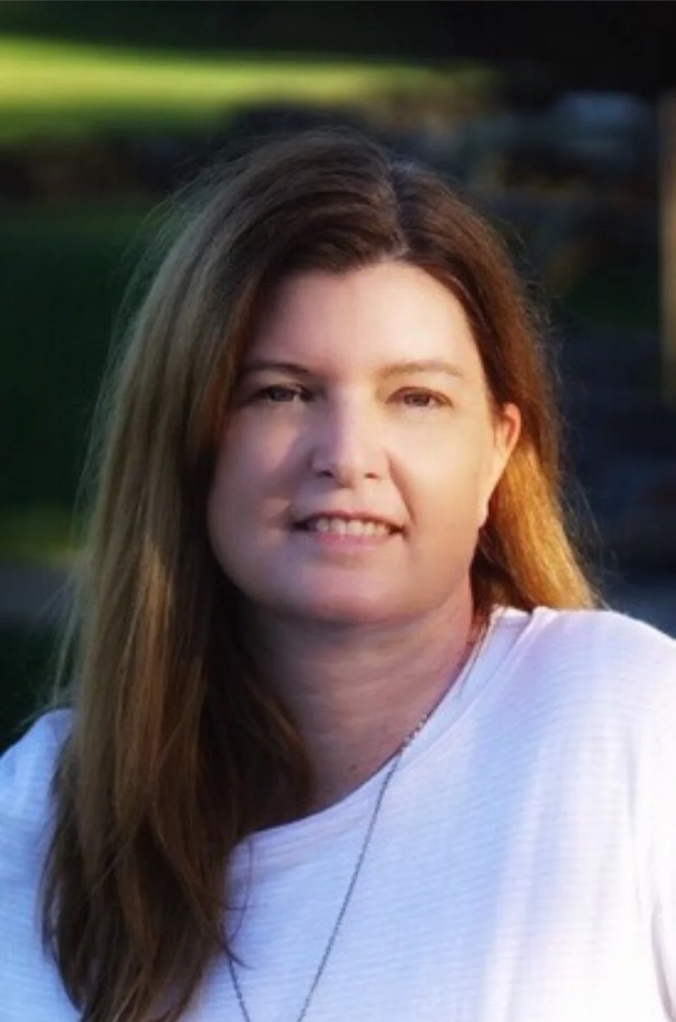 Portrait of a woman with long brown hair, smiling, outdoors during the daytime.
