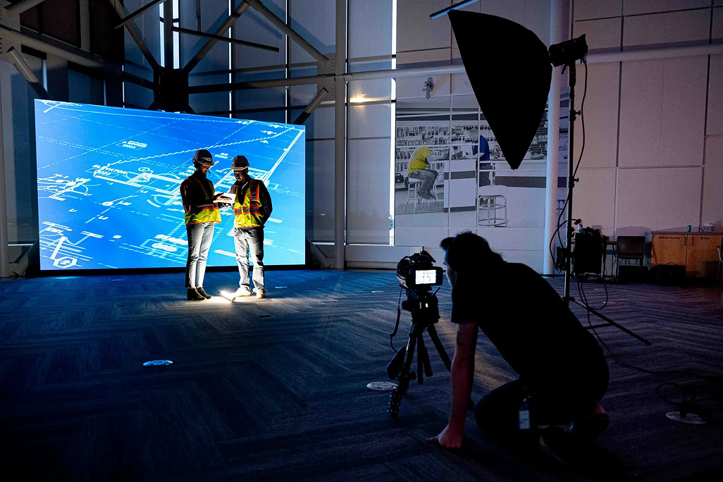 Two people in safety vests and hard hats standing and talking in front of a large digital blueprint display, with a photographer capturing the scene in a studio with professional lighting equipment.