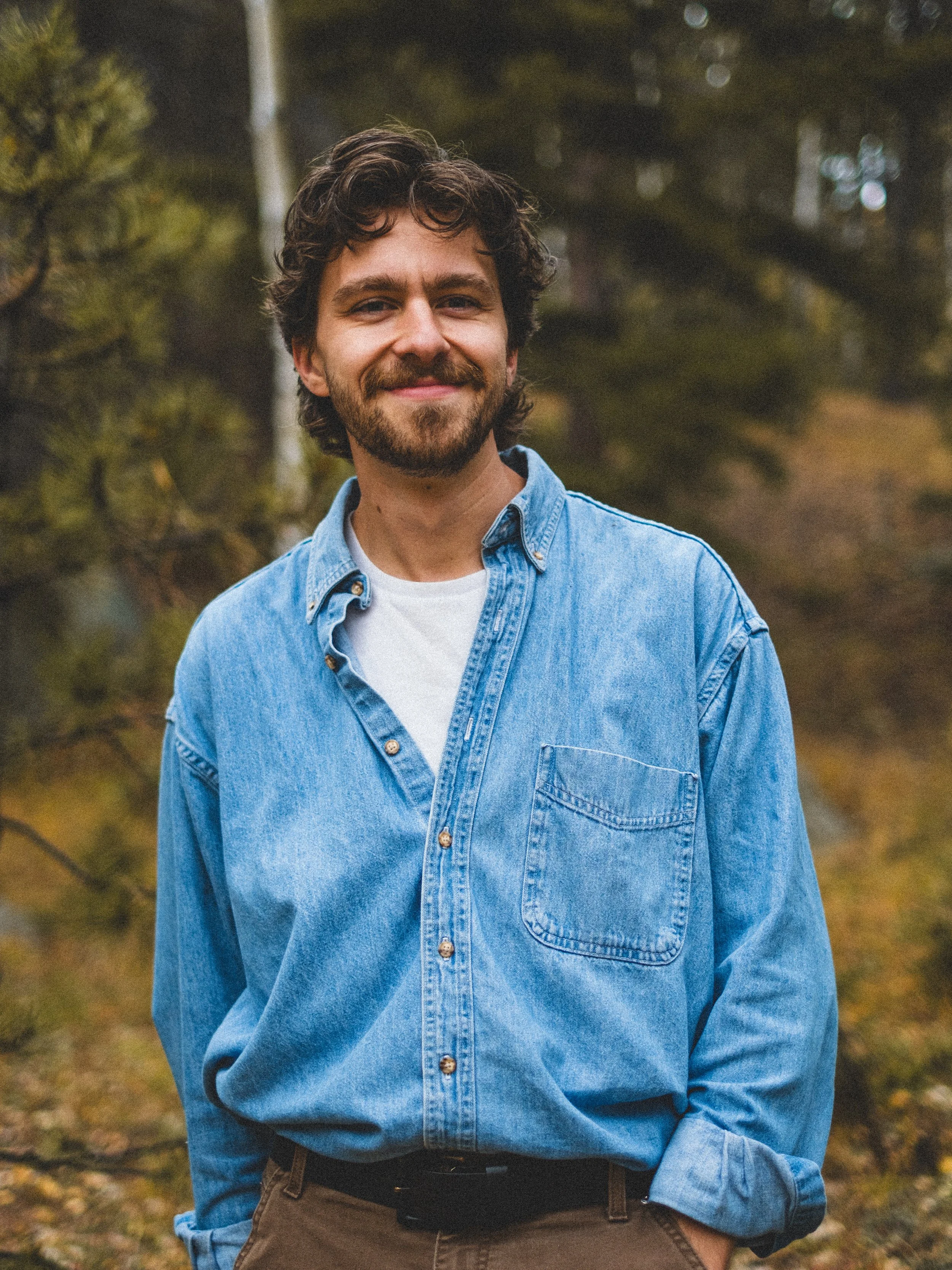 A man with curly hair and a beard standing outdoors in a forest, wearing a denim shirt and smiling at the camera.