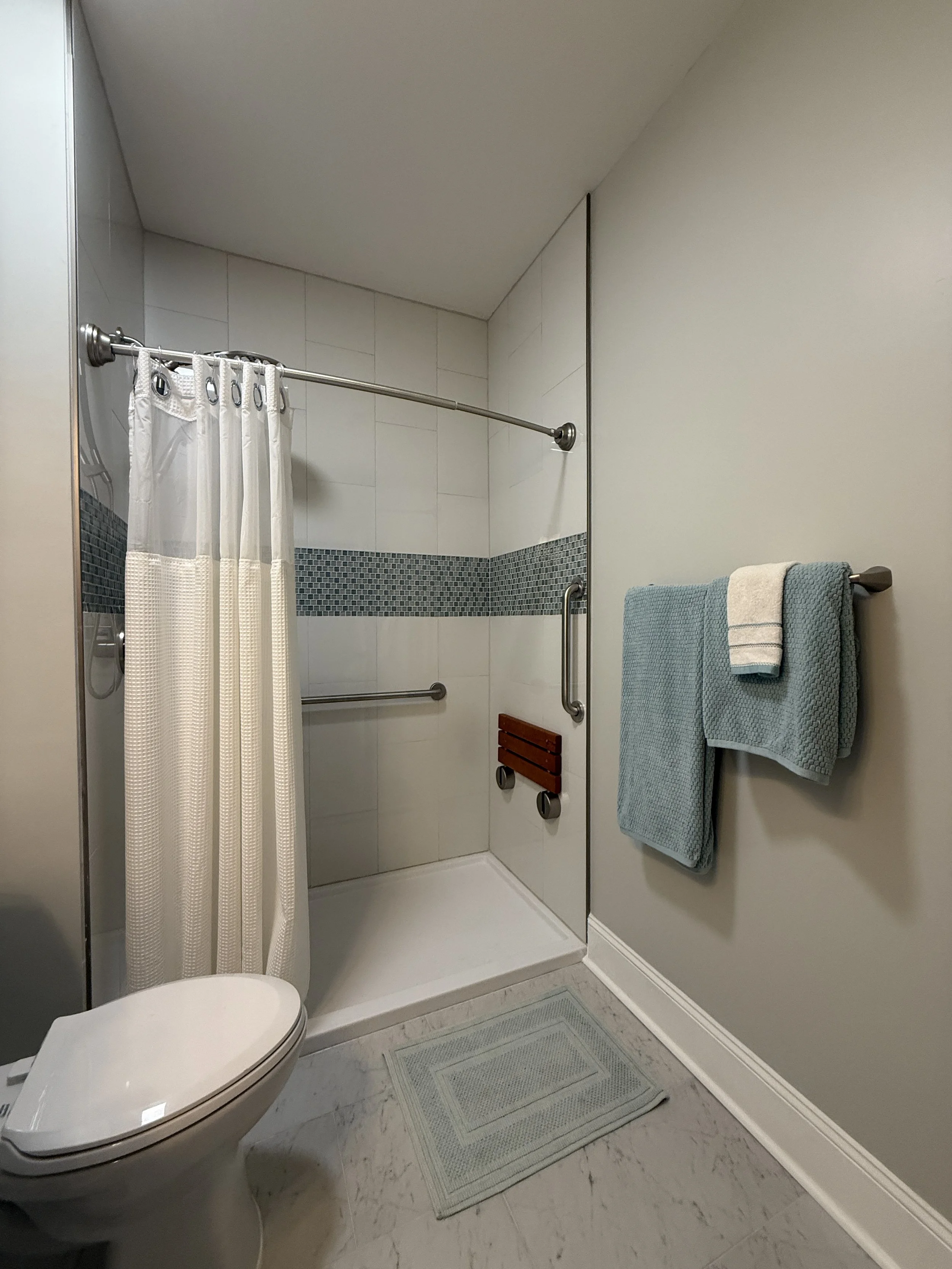 Bathroom shower area with a curtain, gray and white tiles, a grab bar, and a small wooden seat, with towels hanging on the wall.